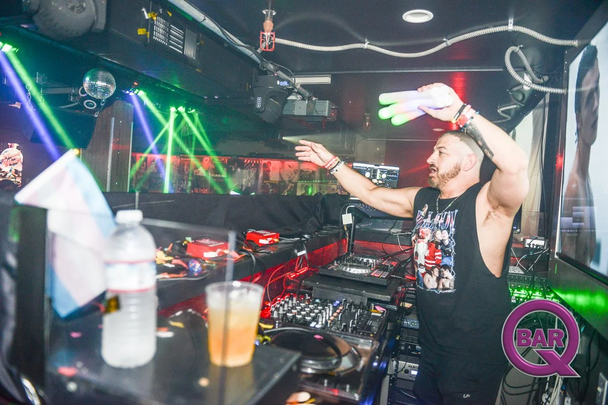 A DJ in a nightclub, wearing a sleeveless shirt, is operating DJ equipment with colorful lights and laser effects in the background, and a purple 'Bar Q' logo in the lower right corner.