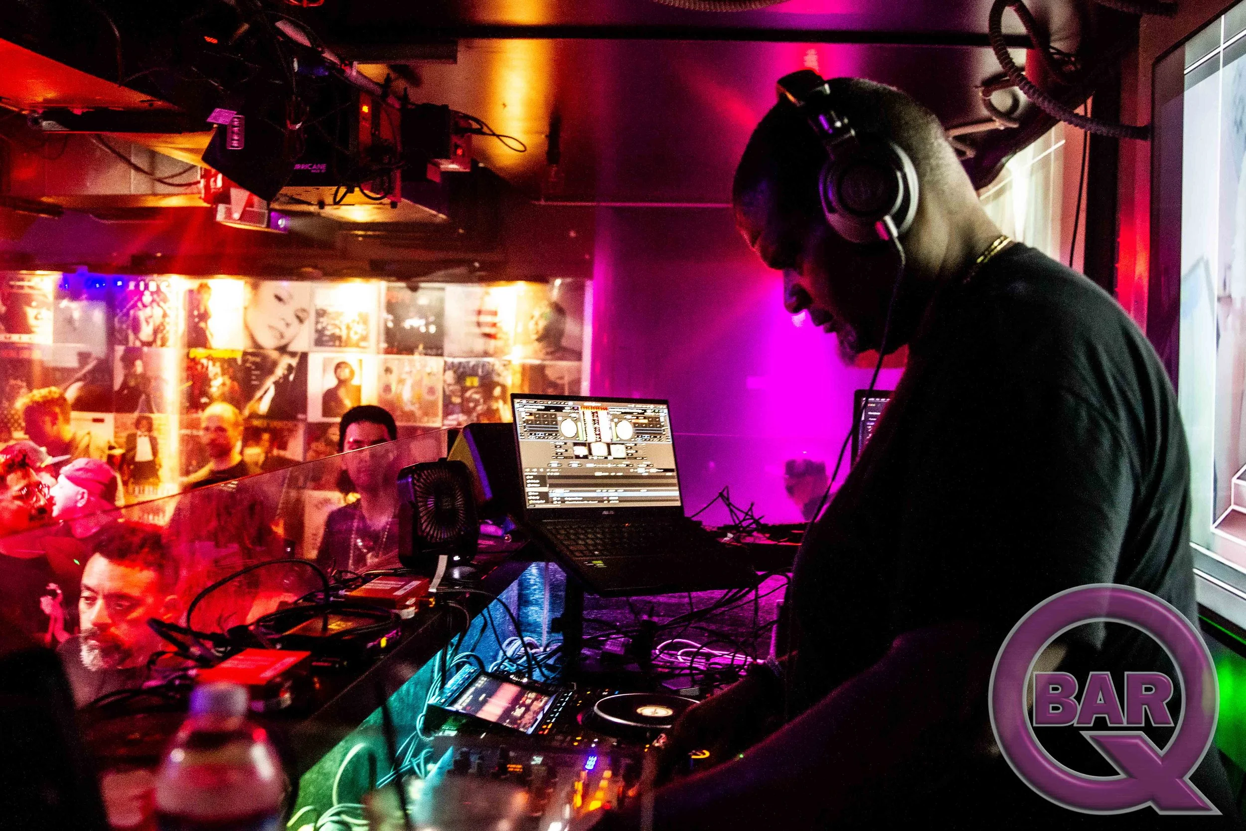 A DJ wearing headphones is mixing music at a nightclub with colorful lights and a crowd of people watching.