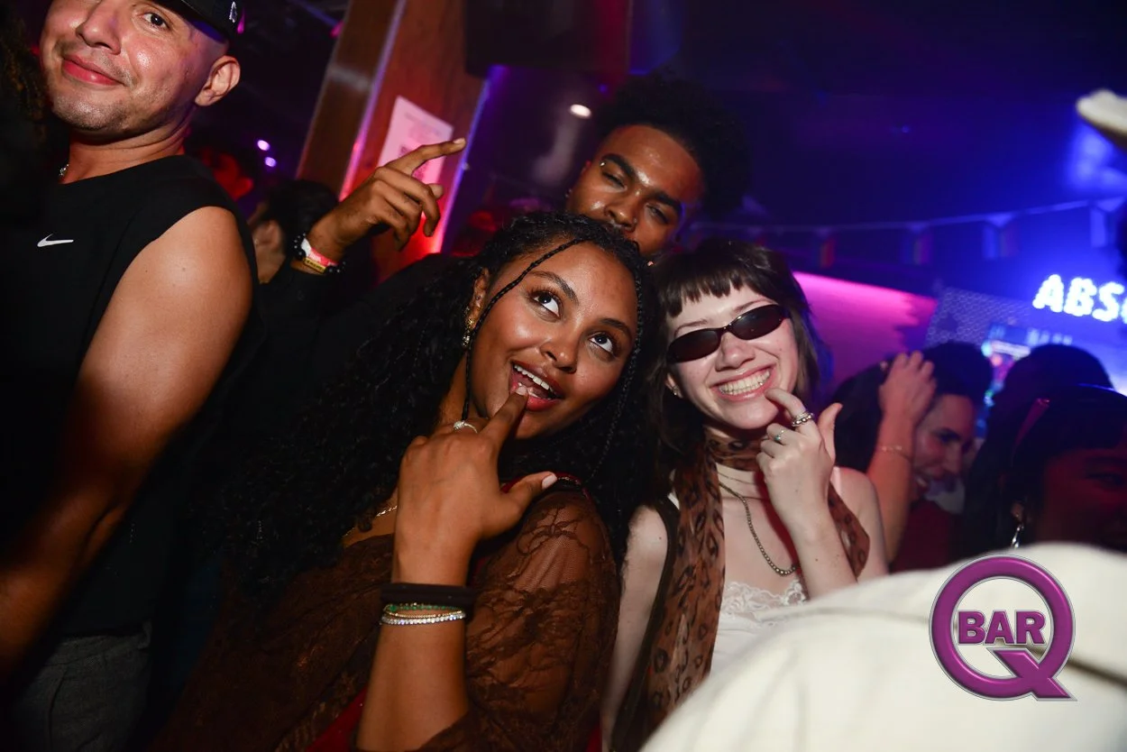 Group of diverse friends partying at a nightclub, smiling and having fun, with colorful lighting and a "BAR" sign in the background.
