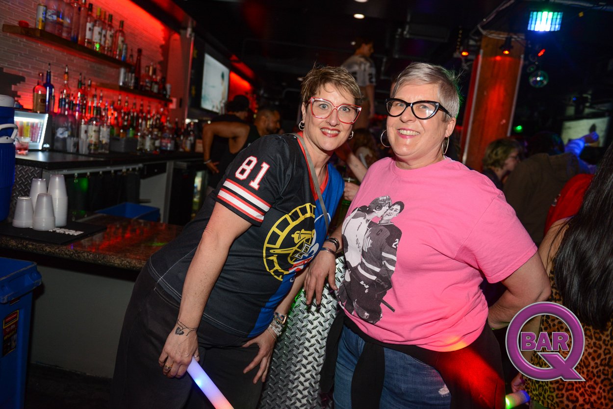 Two women posing and smiling in a bar with colorful lights and a bartender in the background. One woman is wearing a black sports jersey with the number 81 and the other is in a pink T-shirt with hockey players printed on it.