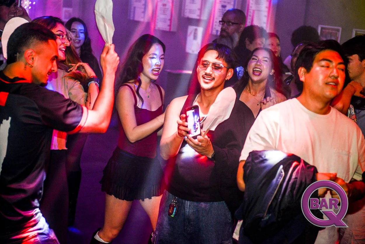 People dancing and having fun at a bar or nightclub, illuminated by colorful lights, with some holding drinks and taking photos.