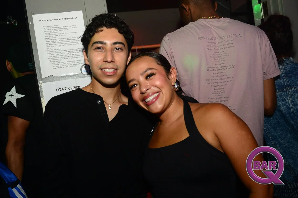 Two smiling people, a young man and a woman, at a dance event, standing close together with their heads touching. The man has dark curly hair and is wearing a black shirt, and the woman has dark hair pulled back, wearing hoops and a black tank top. T