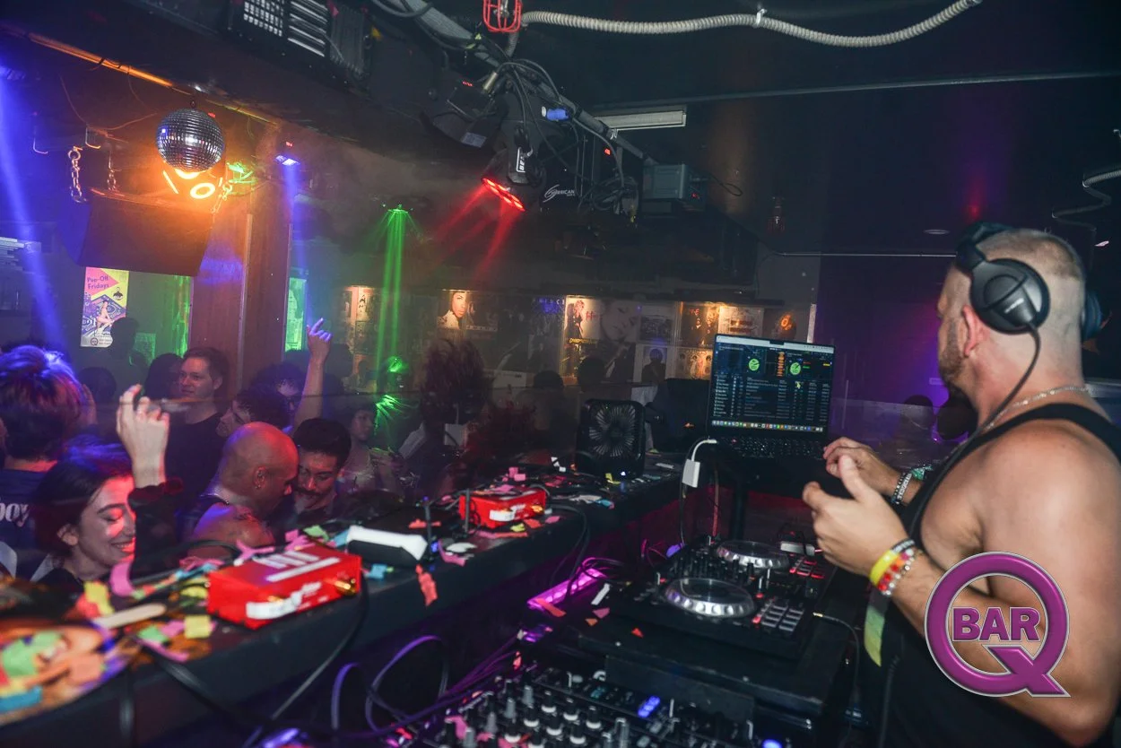 A DJ plays music at a nightclub with a crowd dancing and enjoying the party. The club features colorful lighting, a disco ball, and framed posters on the walls.
