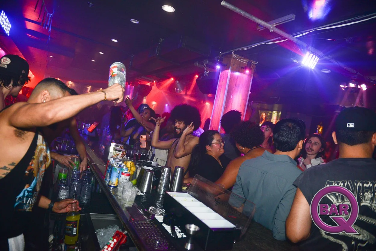 People dancing and socializing at a lively nightclub bar with colorful lights, bottles, and glasses on the counter.