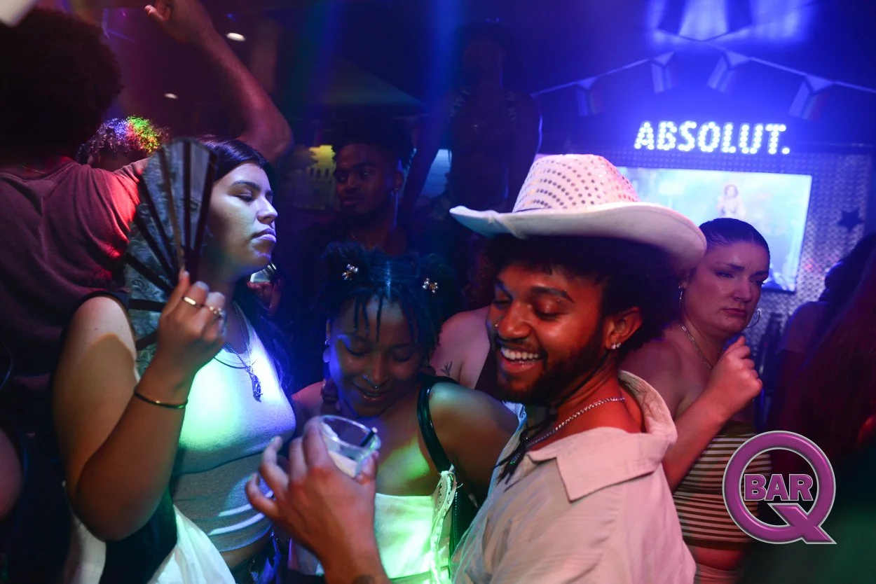 People dancing and socializing at a bar with colorful lighting, including a man wearing a cowboy hat smiling, and a large 'ABSOLUT' sign in the background.