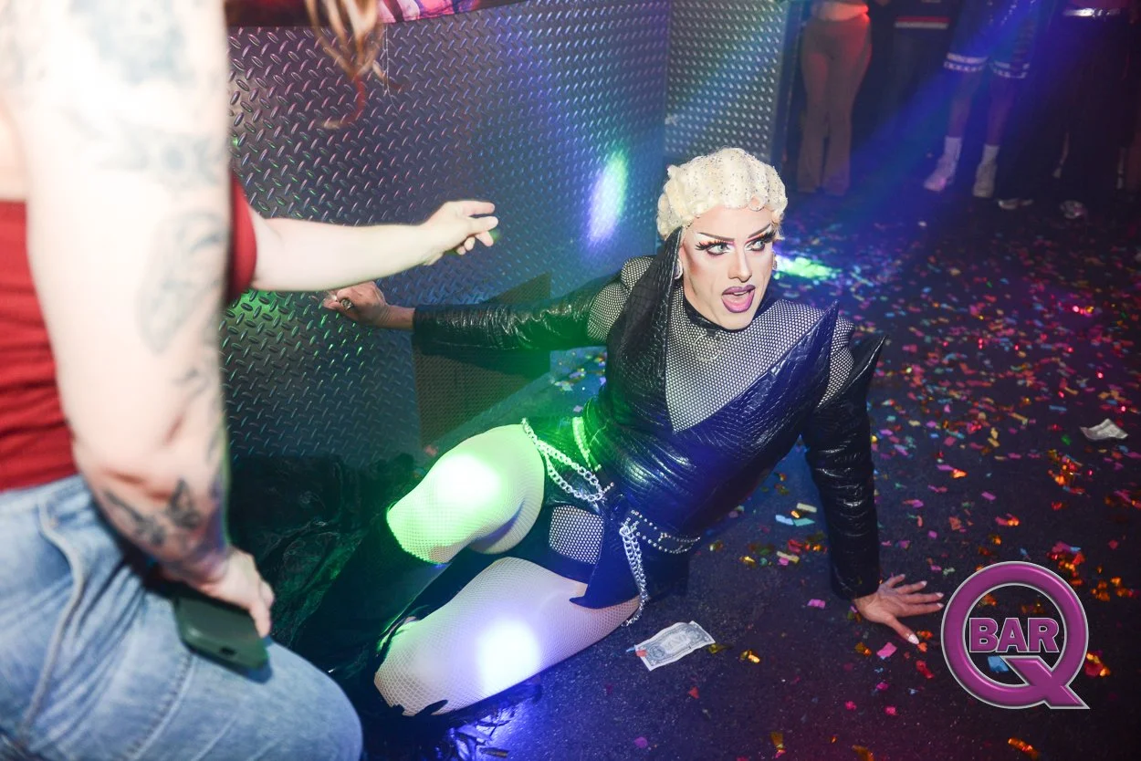 Person with blonde hair, dressed in black, kneeling on club floor, looking surprised or shocked, with confetti scattered around, in a nightclub setting with colorful lights and other people in the background.