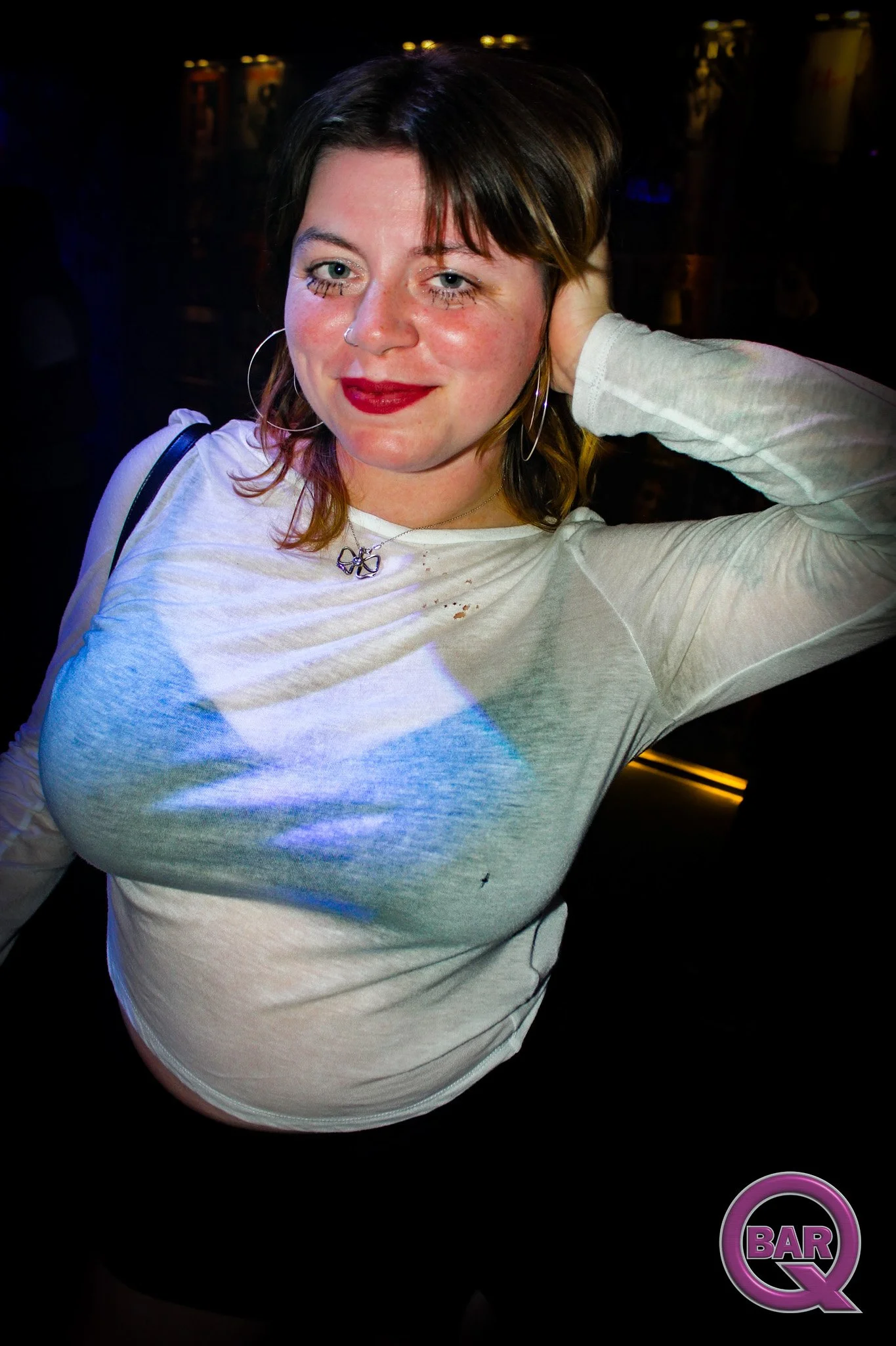 A woman with short brown hair, wearing a white long-sleeve shirt, red lipstick, earrings, and a butterfly necklace, standing in a dark bar or club with a few illuminated cans in the background, and a purple logo with the word "BAR" and a Q at the bot
