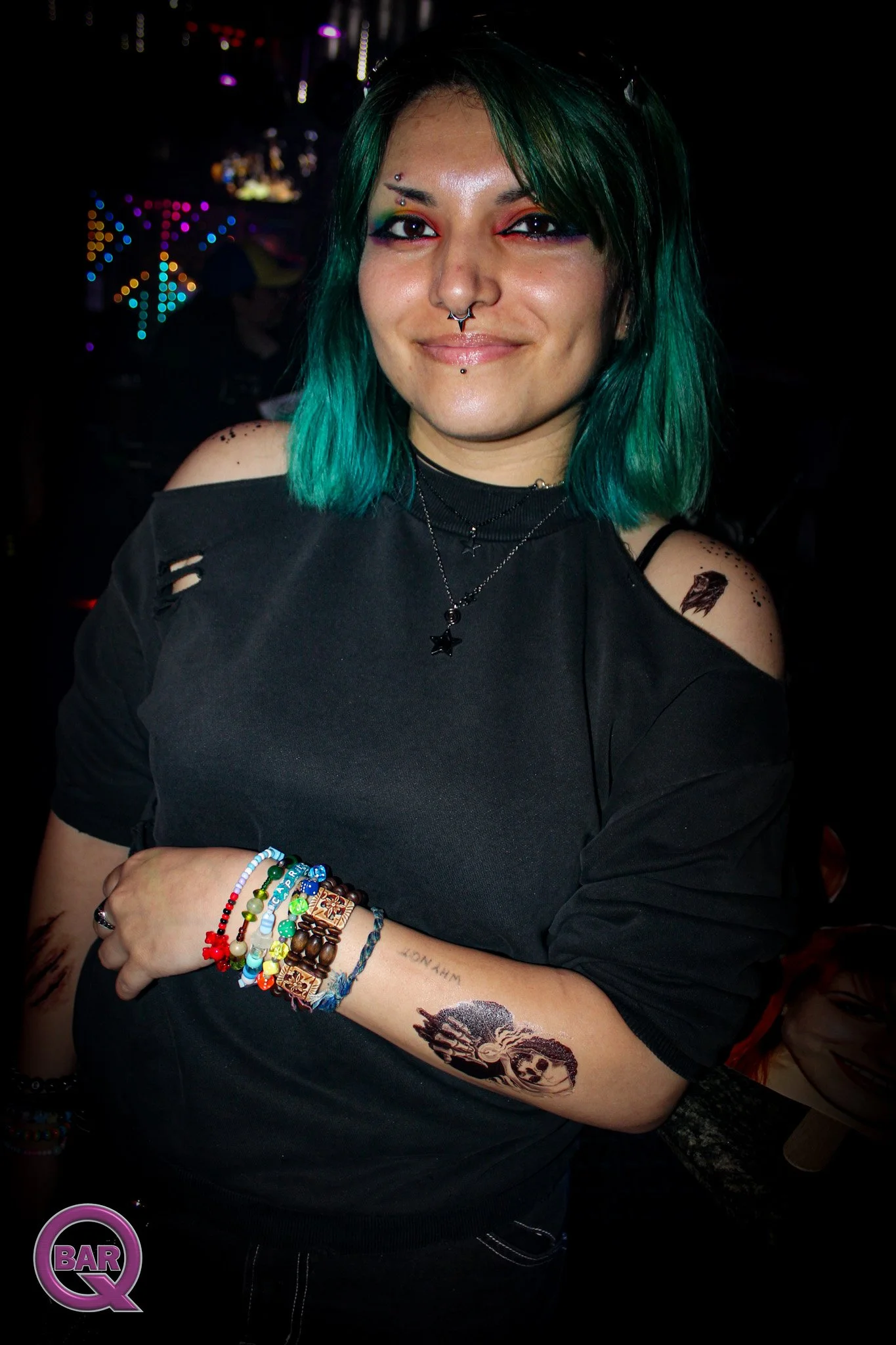 A young woman with teal hair and multiple facial piercings, wearing a black off-shoulder top with ripped sleeves, stands in a dimly lit setting with colorful lights. She has tattoos on her left arm and is wearing multiple colorful bracelets. She has 