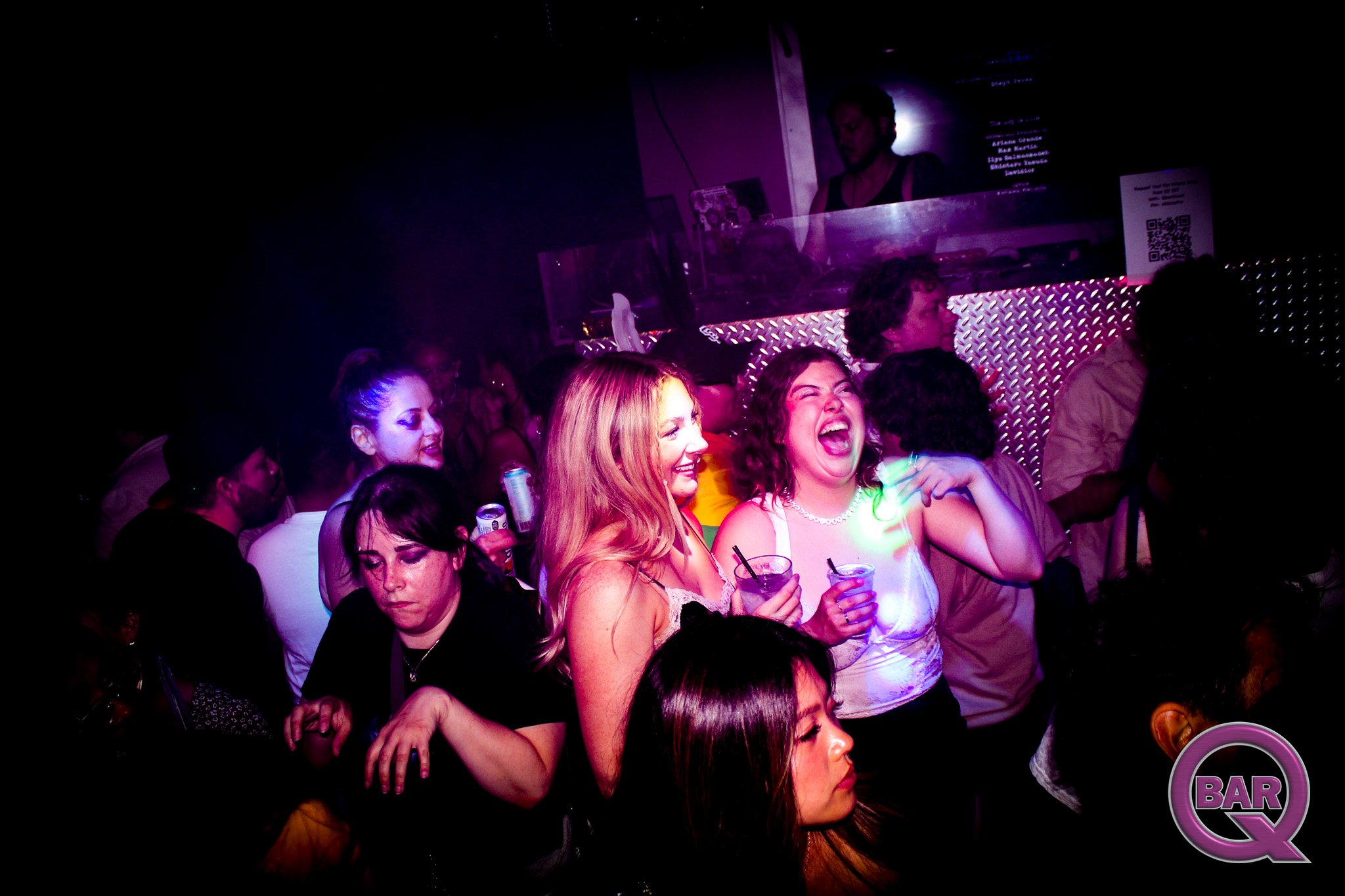 Group of people at a nightclub dancing and enjoying drinks with purple and pink lighting, a DJ is visible behind the bar, and a neon sign with the words 'BAR' is in the background.
