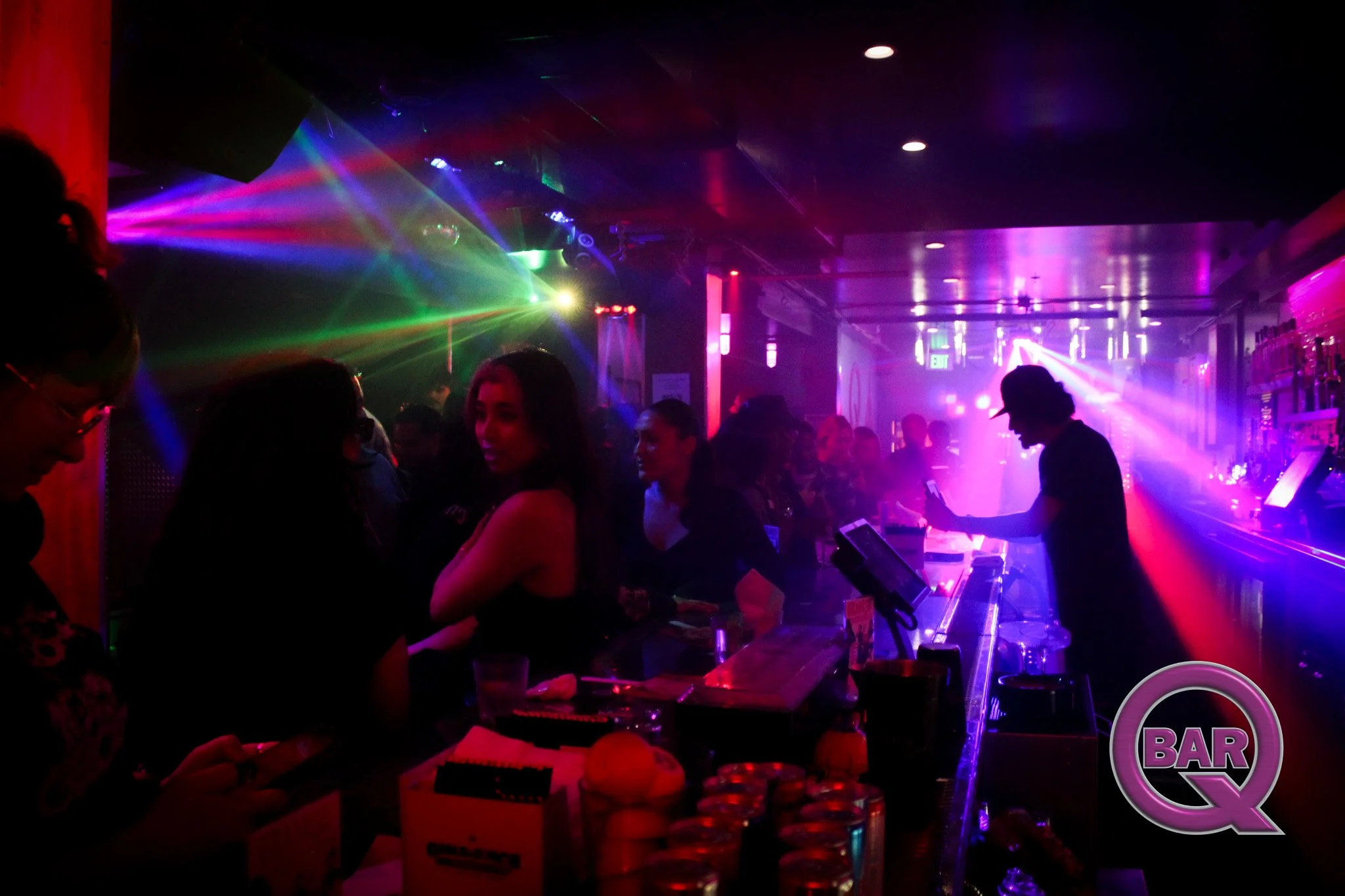 A dimly lit nightclub scene with colorful laser lights, people standing at a bar, and a DJ playing music.