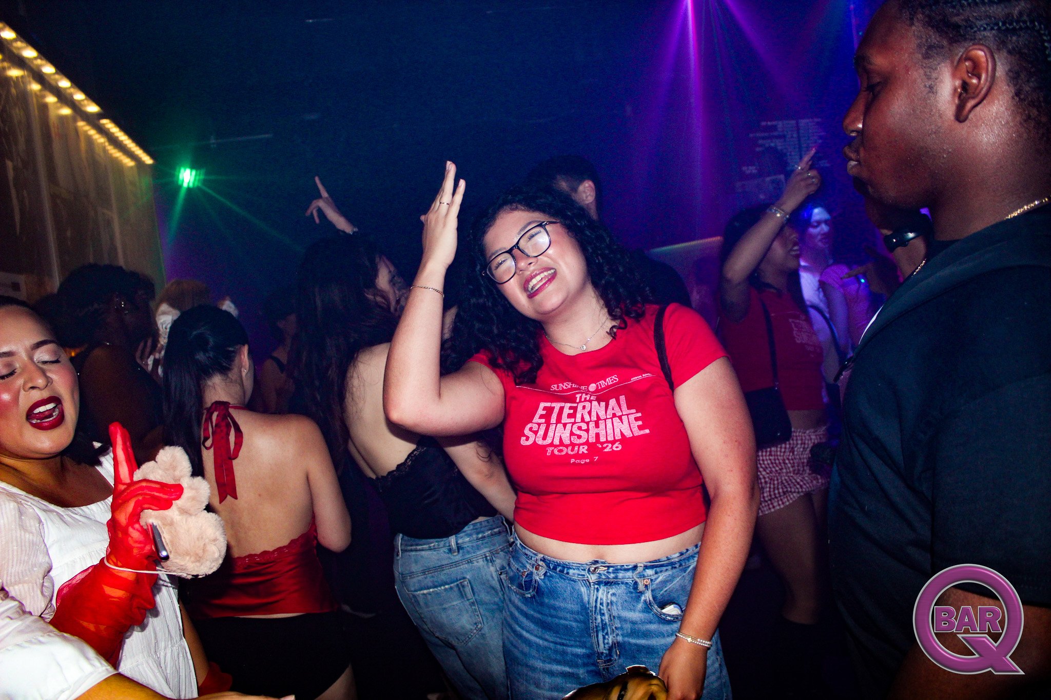 A group of people dancing in a nightclub, with a woman in a red shirt and glasses smiling at the camera, surrounded by others enjoying the music and lights.