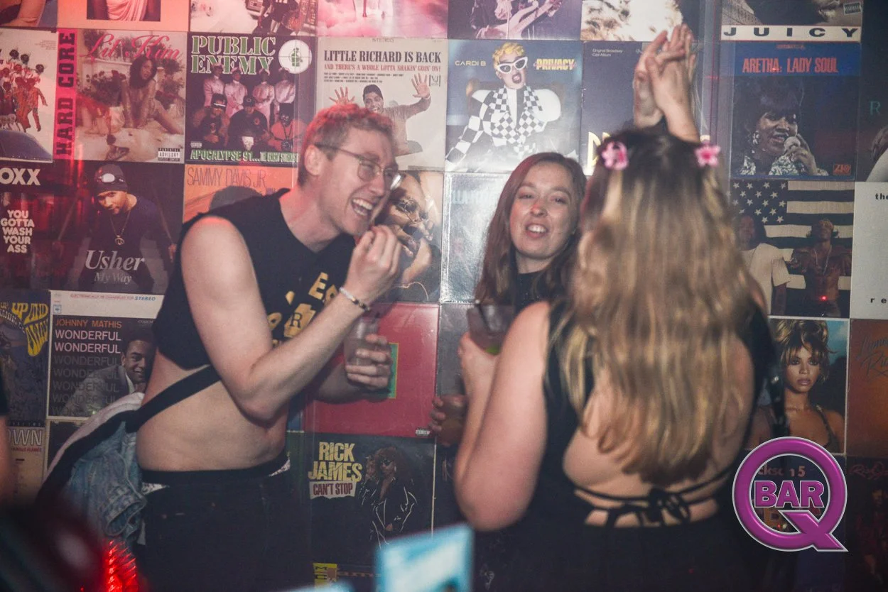 Three people at a bar, two women and one man, smiling and laughing, standing near a wall covered with colorful album covers and posters, with a purple neon Q logo at the bottom right.