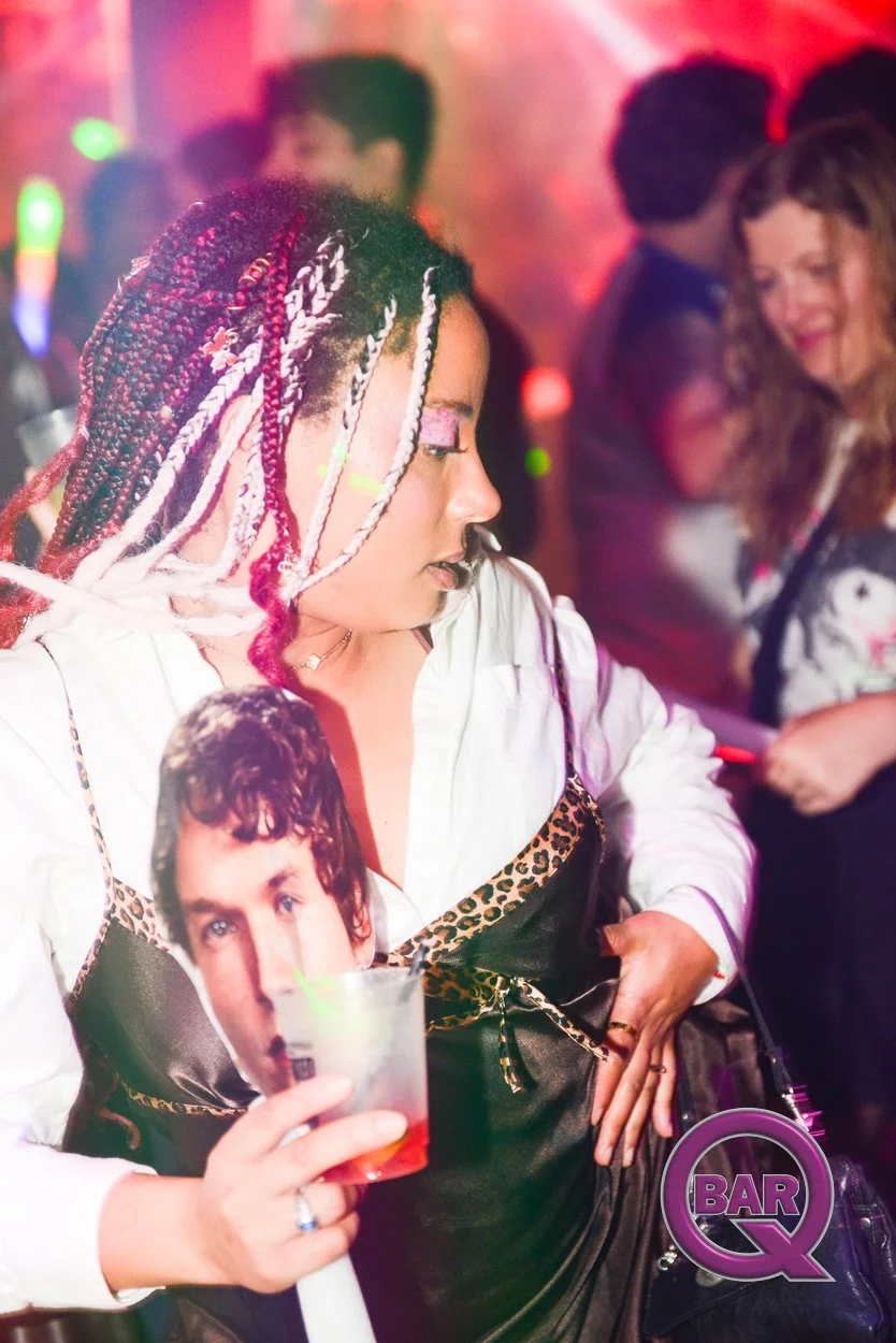 A woman with braided hair and eye makeup holding a drink at a lively party or nightclub. She is wearing a shirt with a large print of a man's face. Several people are dancing or socializing in the background with colorful lights.