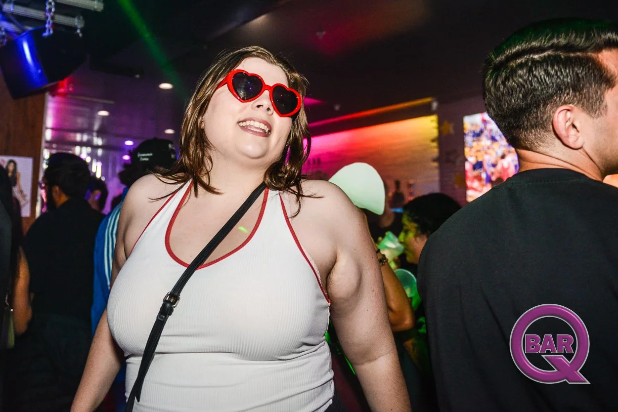 A woman wearing heart-shaped red sunglasses and a white tank top with red trim, smiling and dancing in a nightclub with colorful lighting and a crowd in the background.