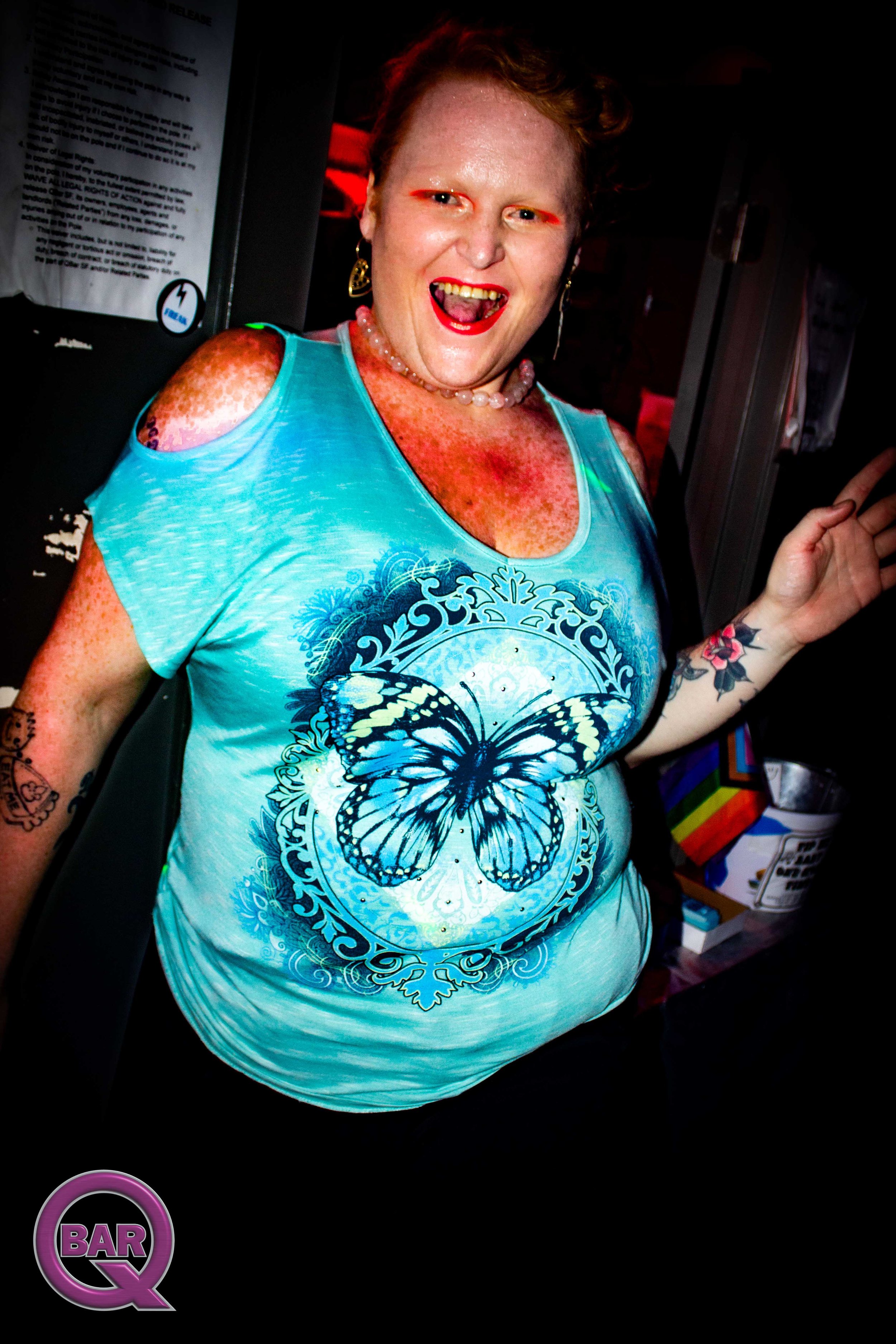 A woman with short, curly red hair, wearing a light blue sleeveless top with a large butterfly design on it, red lipstick, earrings, and a pearl necklace. She is smiling, showing her teeth, and holding up her right hand with two fingers in a peace si