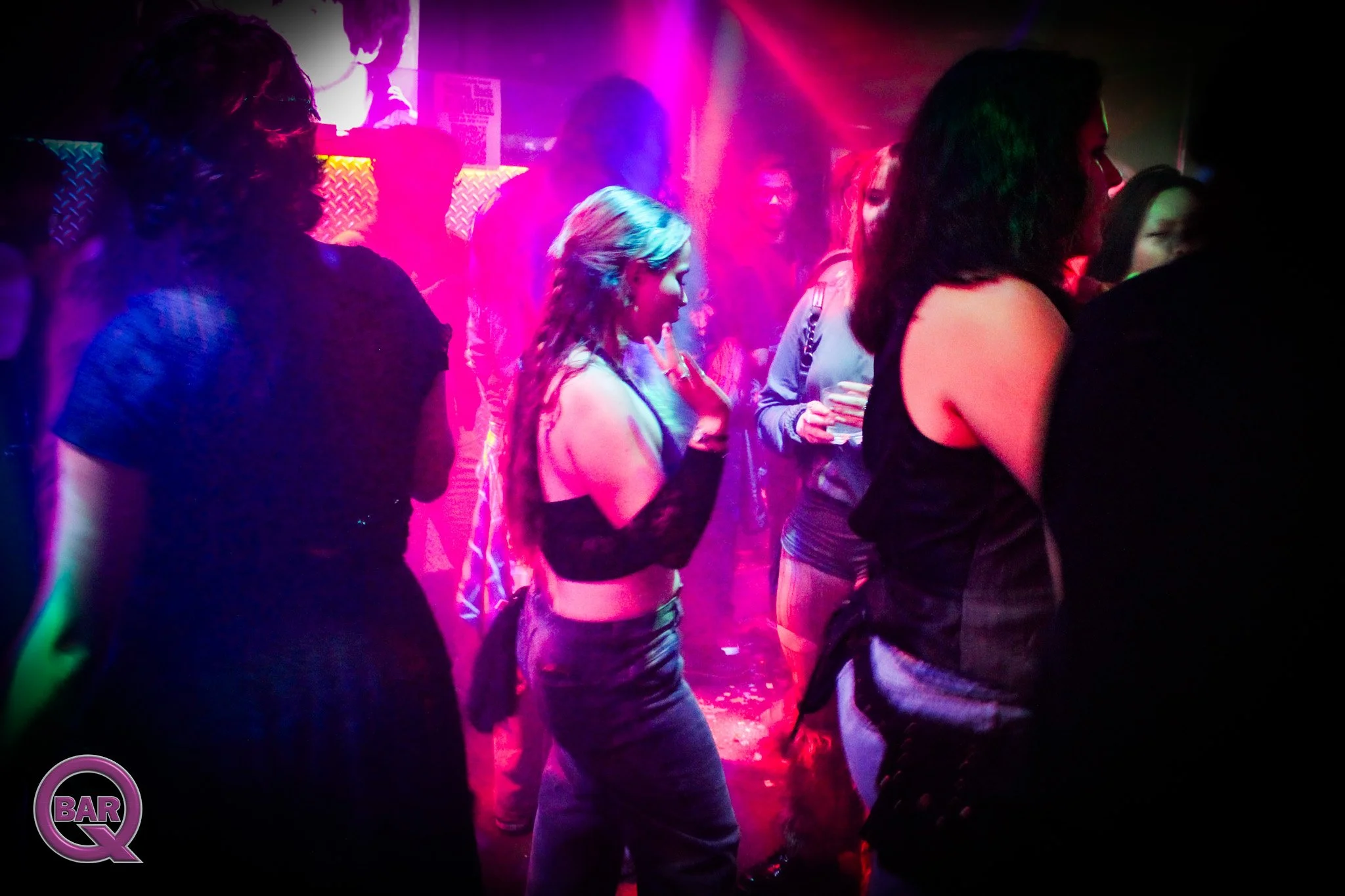 A group of people dancing in a nightclub with colorful purple and pink lighting and a small bar area in the background.