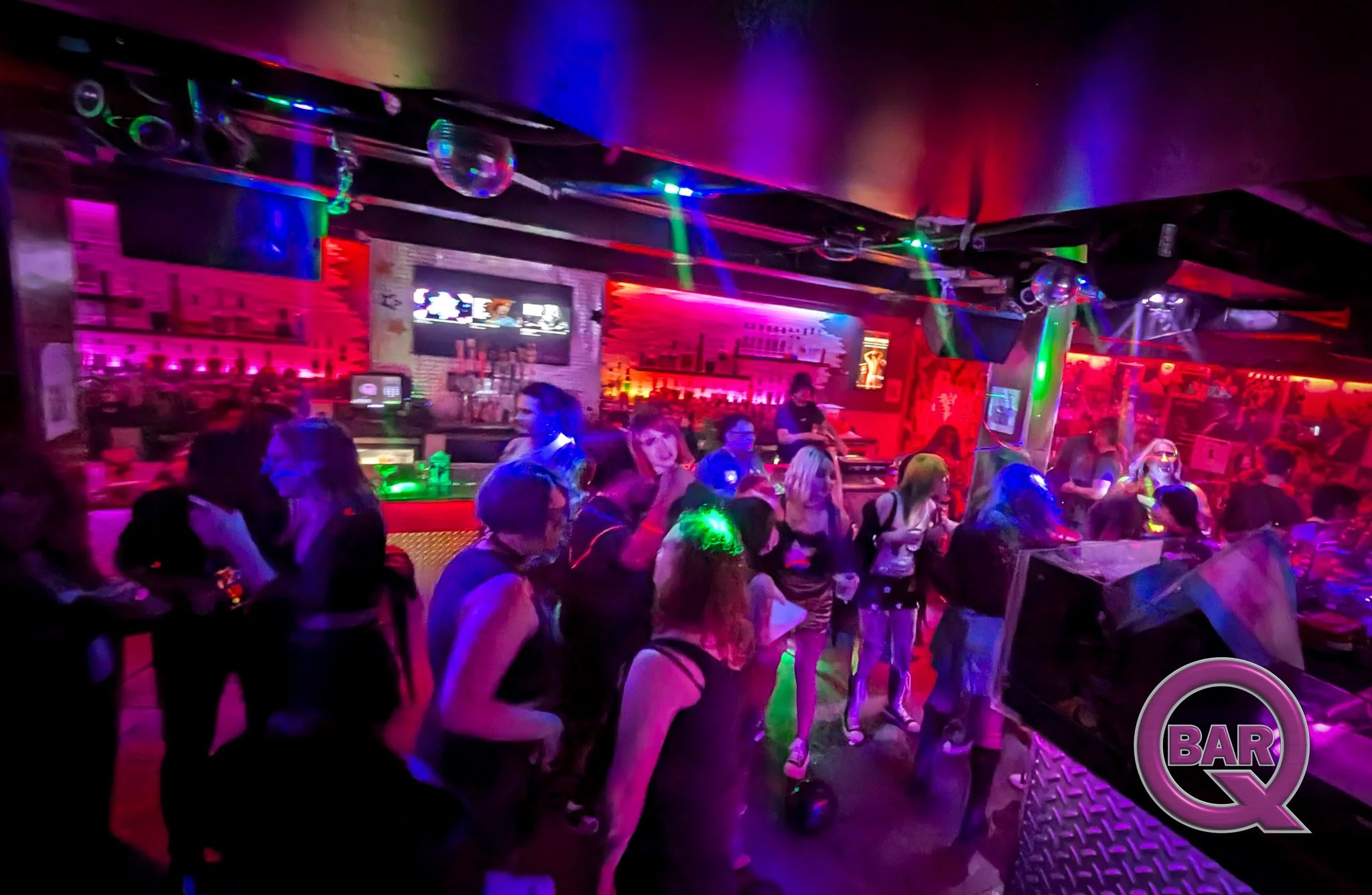 People gathered inside a bar with colorful lights, a large screen, and a neon bar sign, creating a lively atmosphere.