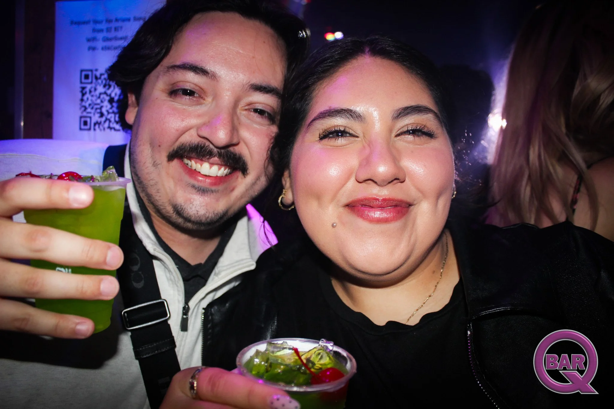 A man and woman smiling at a bar, holding a green cocktail with a cherry and garnish, in a lively setting with colorful lighting.
