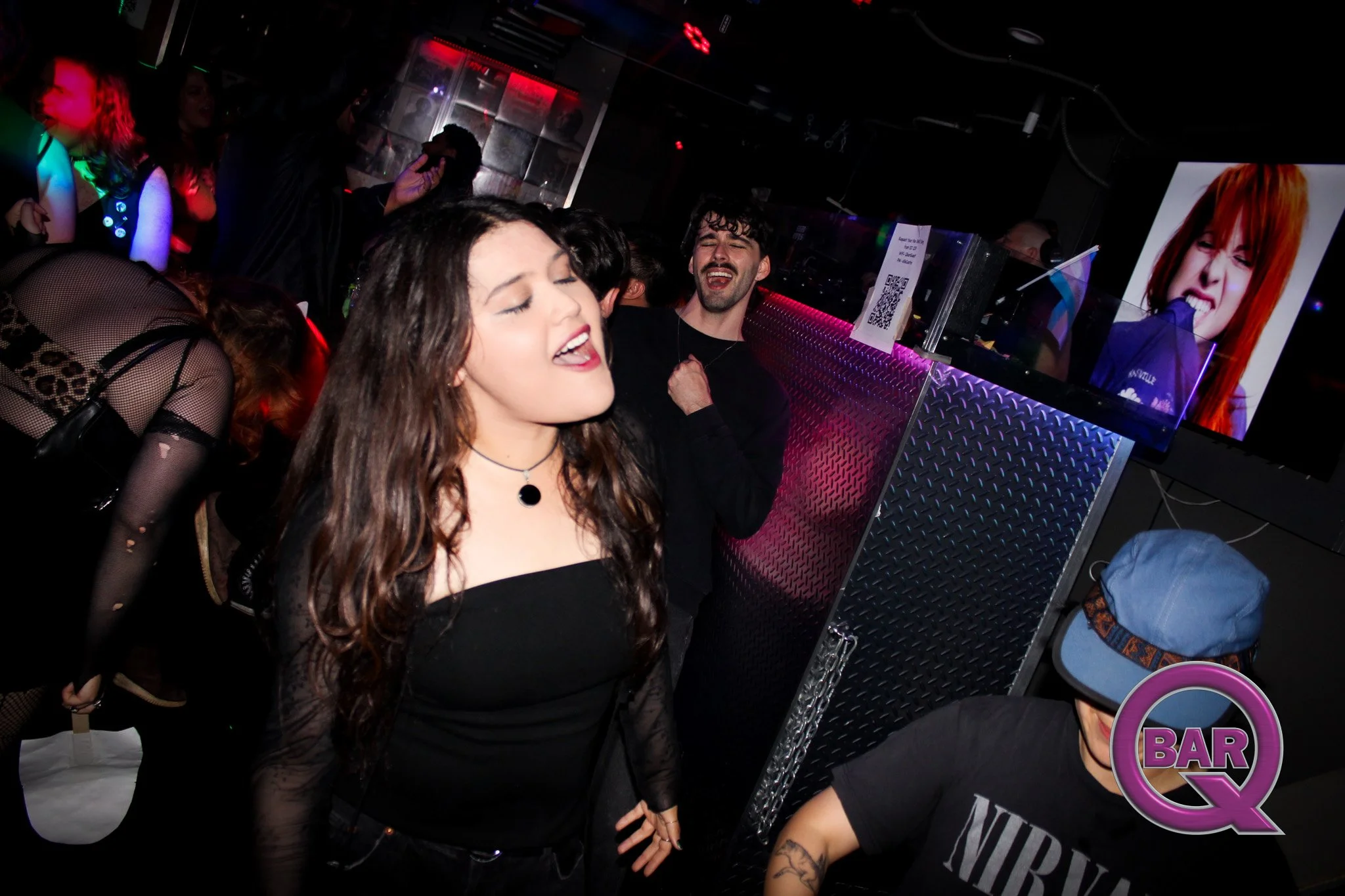 People dancing and enjoying music in a nightclub with colorful lighting and a poster of a smiling woman with red hair.
