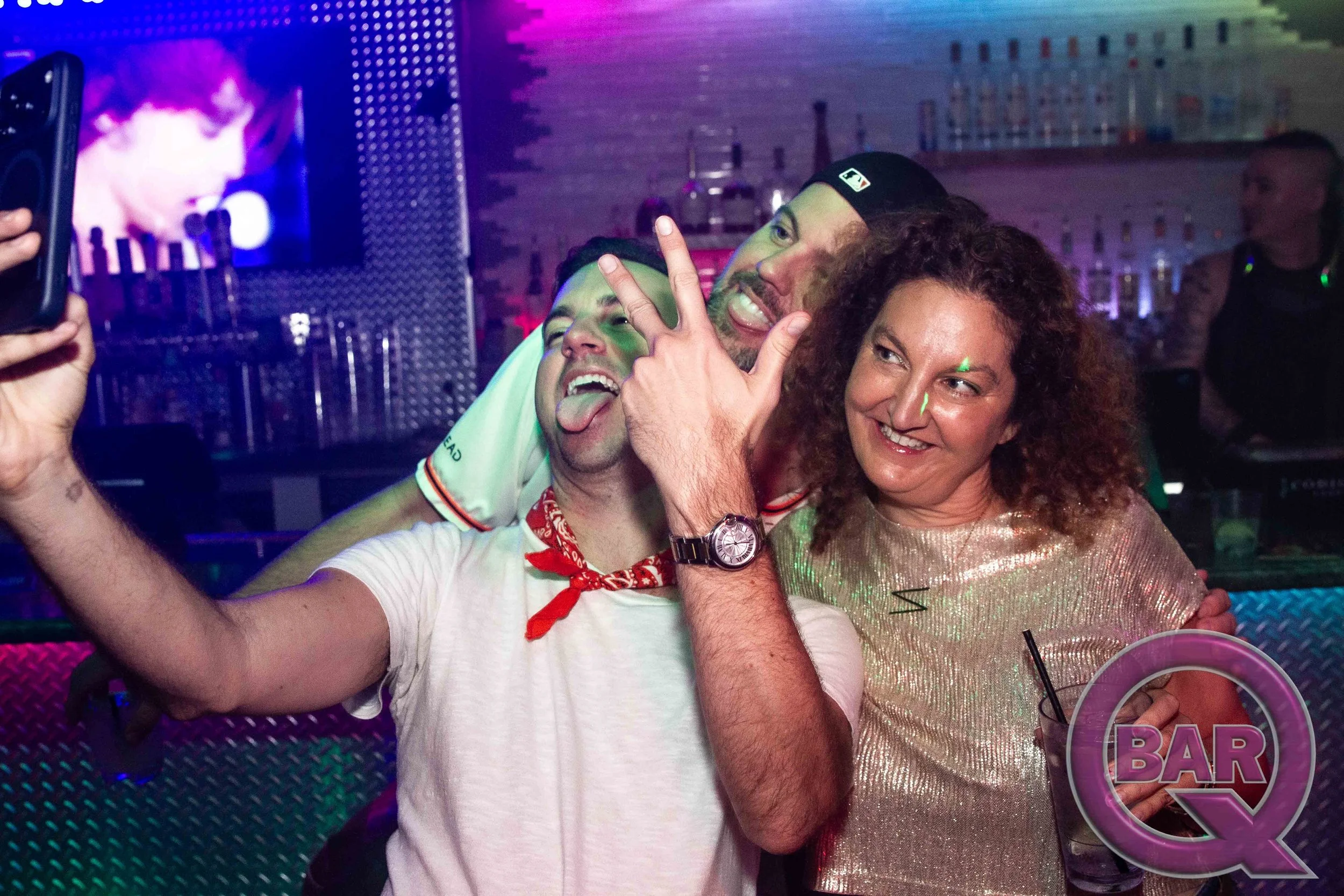 Three people taking a selfie at a bar with colorful lighting. One man in a white shirt makes a peace sign and sticks out his tongue; a woman with curly hair smiles; another man in a black cap is smiling.