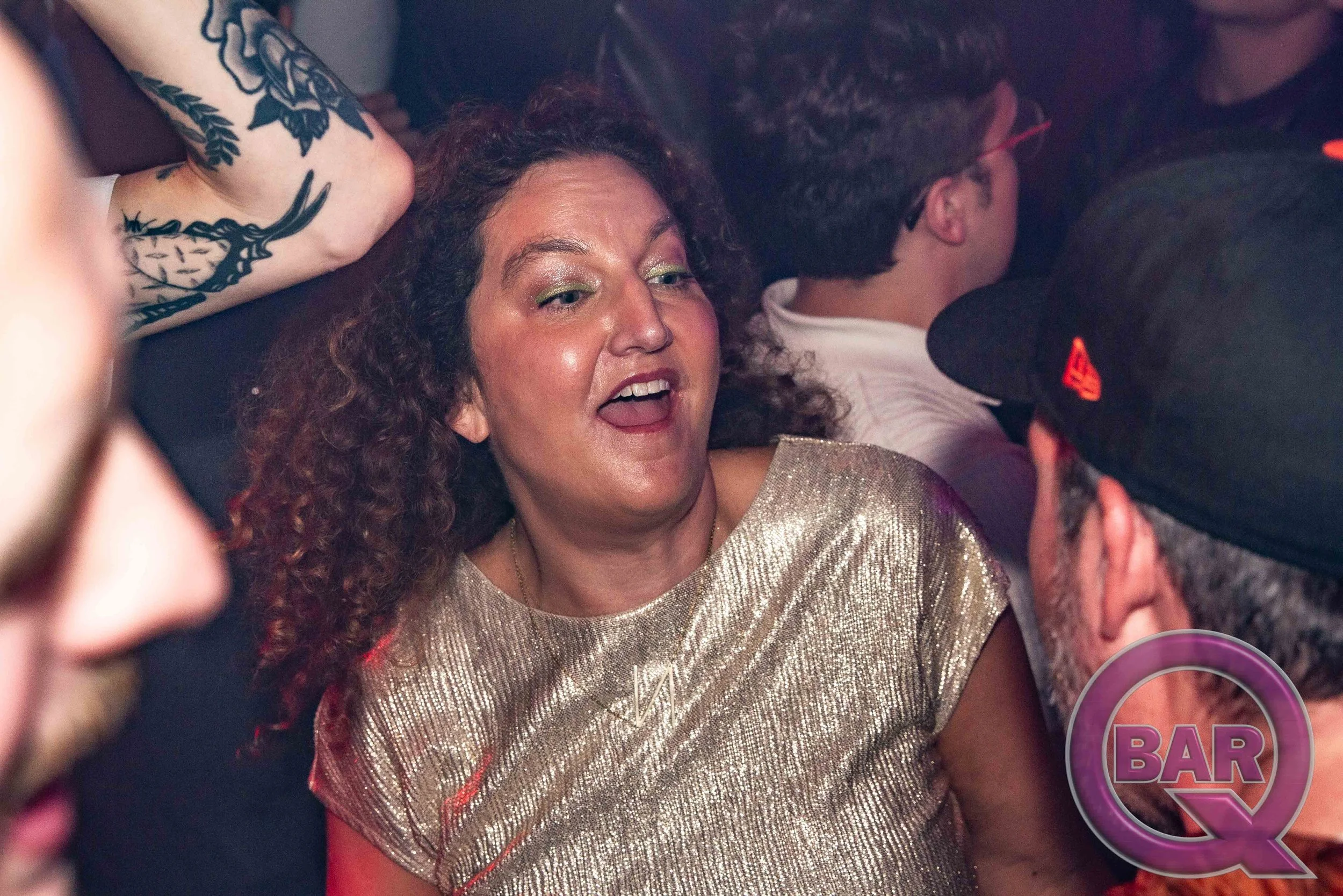 A woman with curly hair and makeup wearing a shiny gold dress, smiling and talking to a man wearing glasses and a cap at a social event.