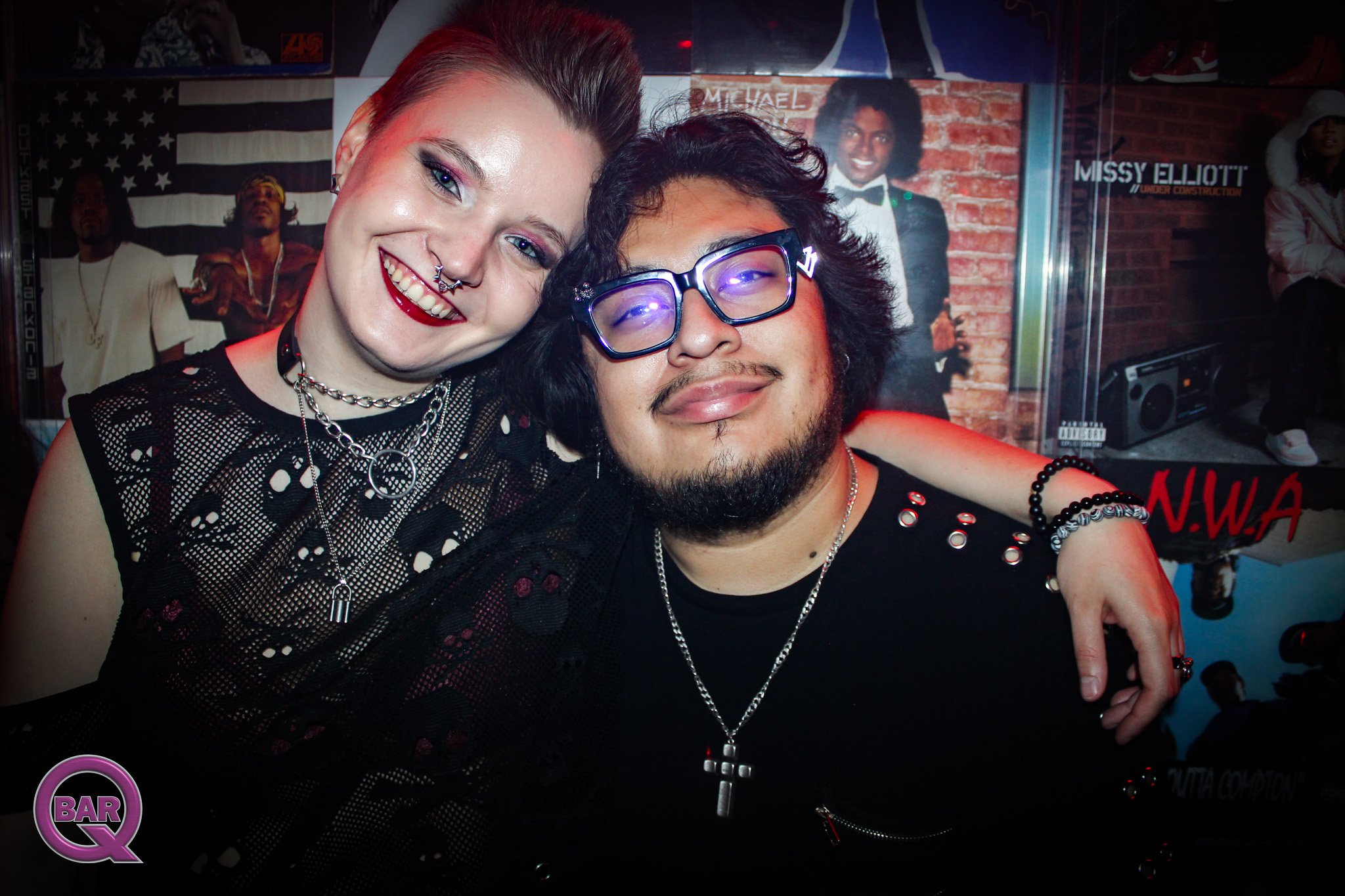 Two smiling young adults, a woman with dyed hair, bright makeup, and accessories, and a man with glasses and a beard, pose closely together at a party or nightclub with posters and people in the background.