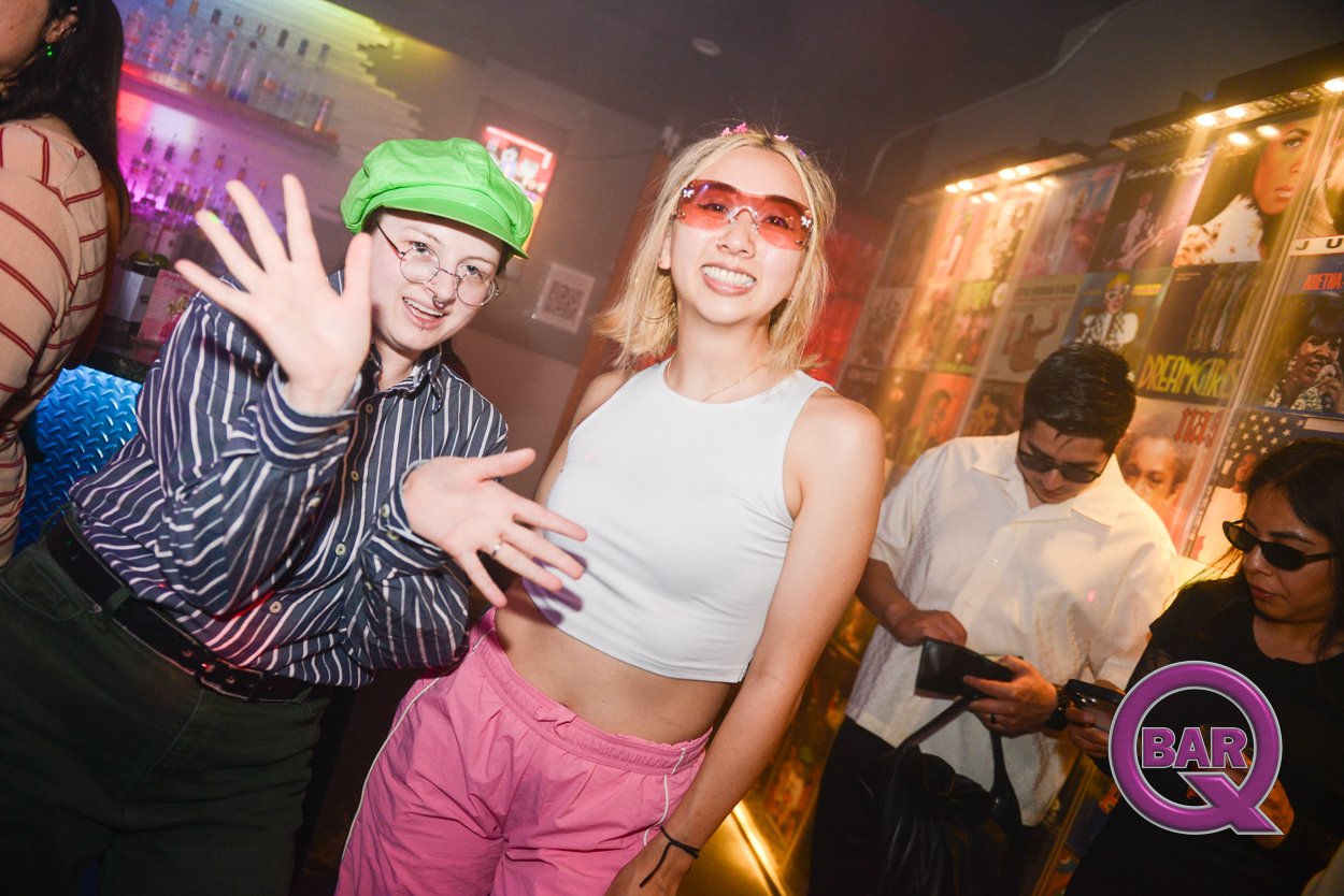 Two women smiling and posing for the camera in a bar, with colorful lighting and posters on the wall behind them; other people in the background looking at their phones.