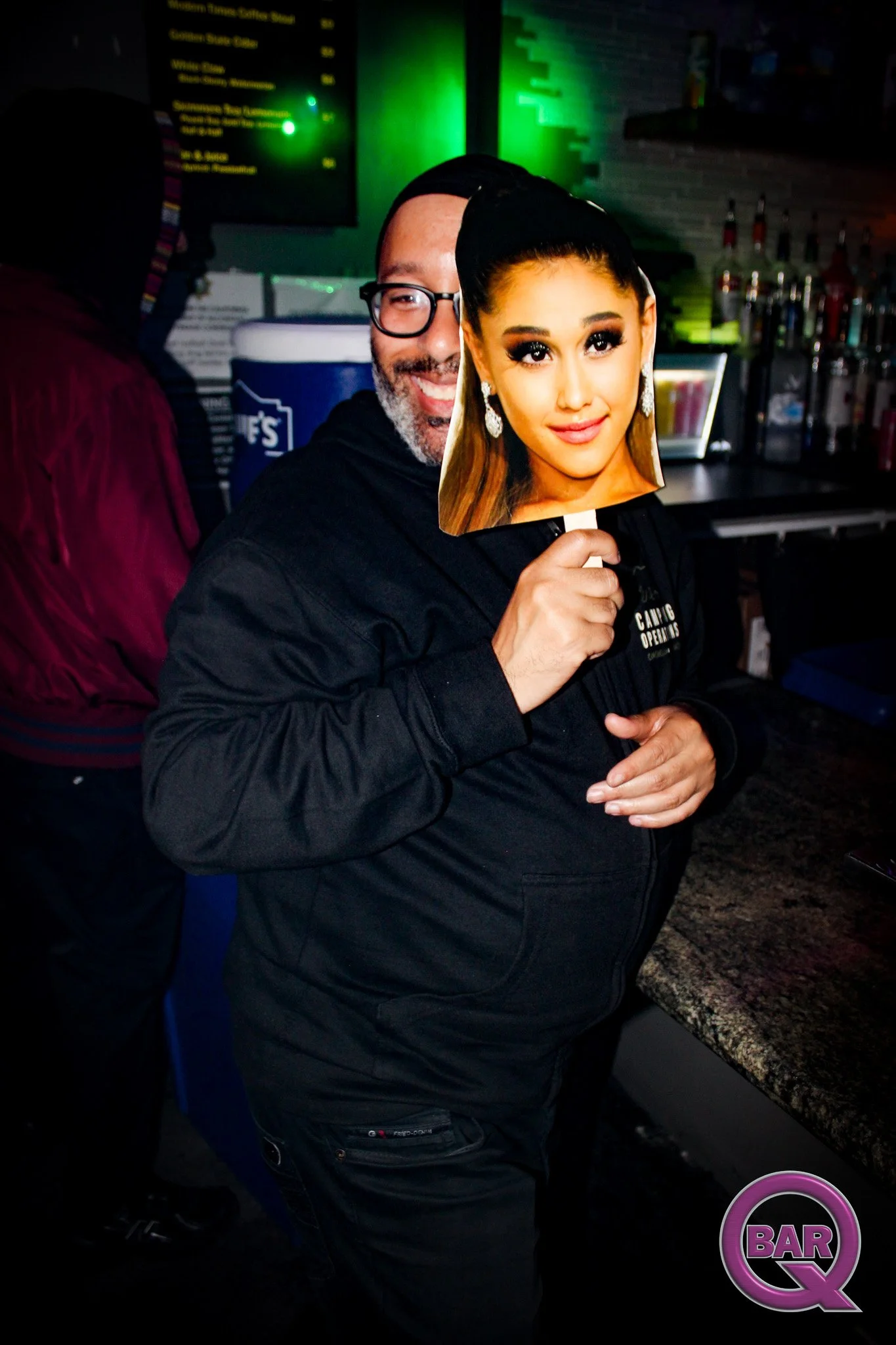 A man holding a cutout of a woman's face mask in a bar or nightclub, with bar-themed signage and bottles in the background.