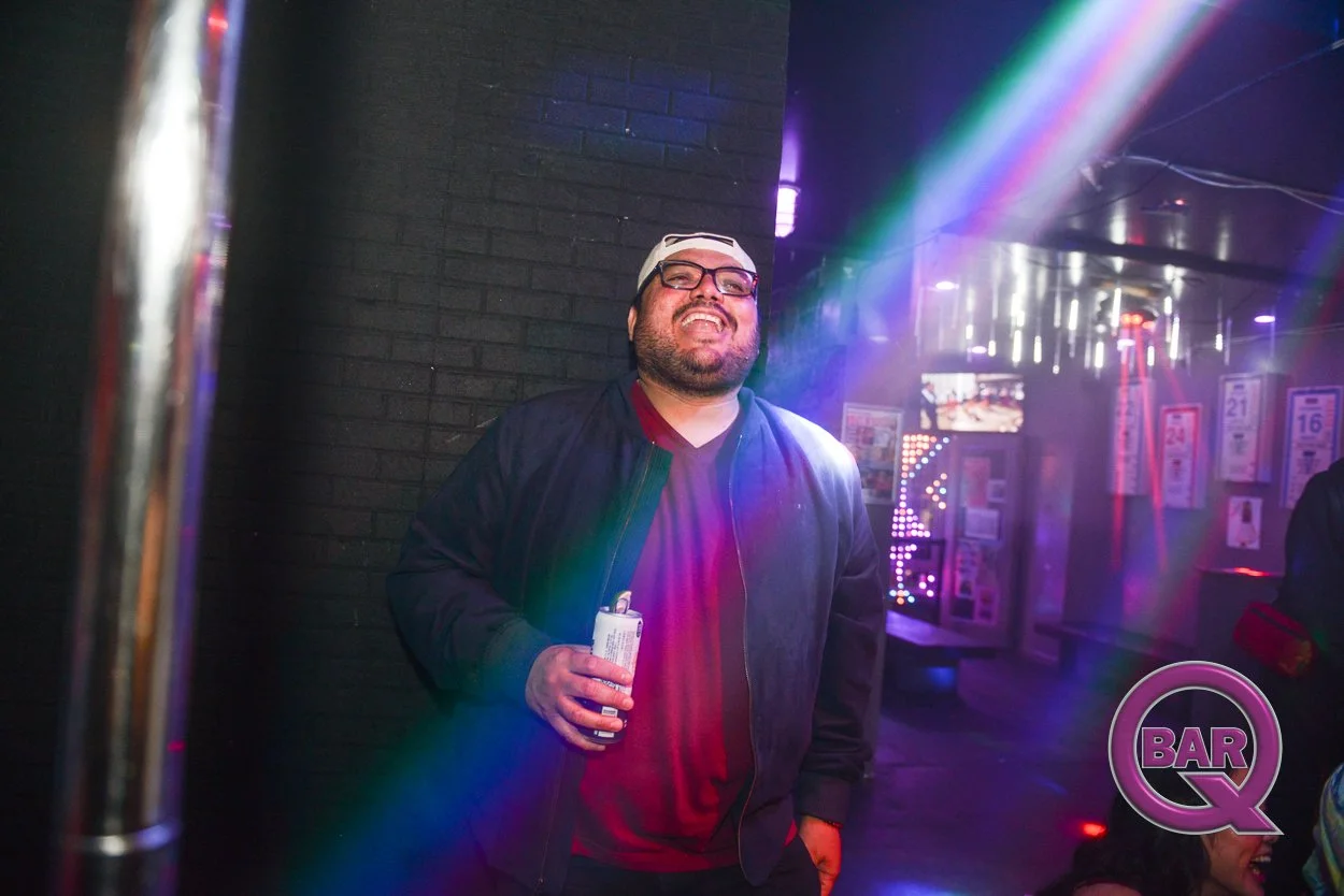 A man with glasses and a beard, wearing a black jacket and red shirt, smiling and holding a bottle, in a dimly lit bar with colorful rainbow light effects.
