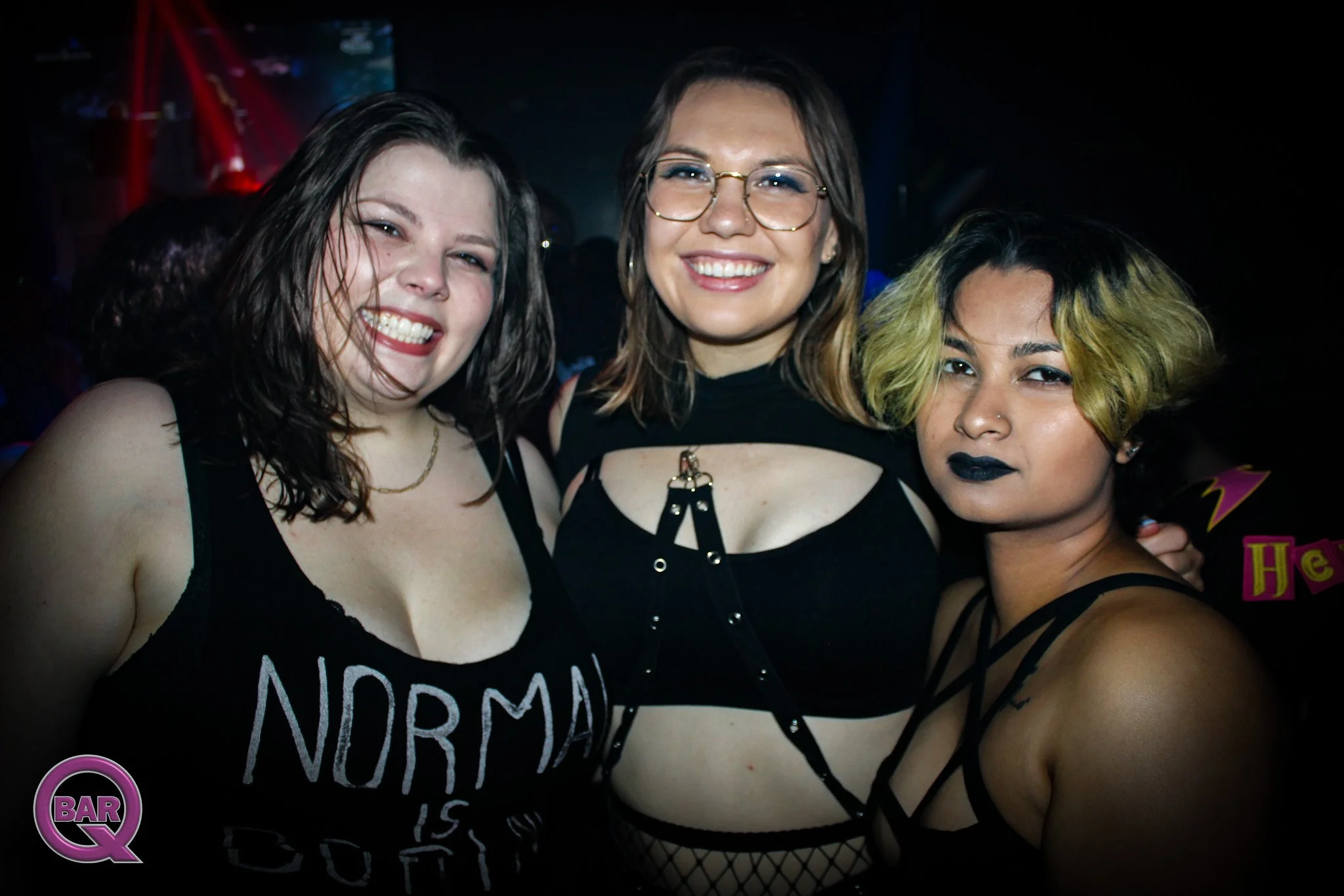 Three women smiling and posing close together in a dark nightclub environment, with colorful lighting and a 'Bar Q' logo in the bottom corner.
