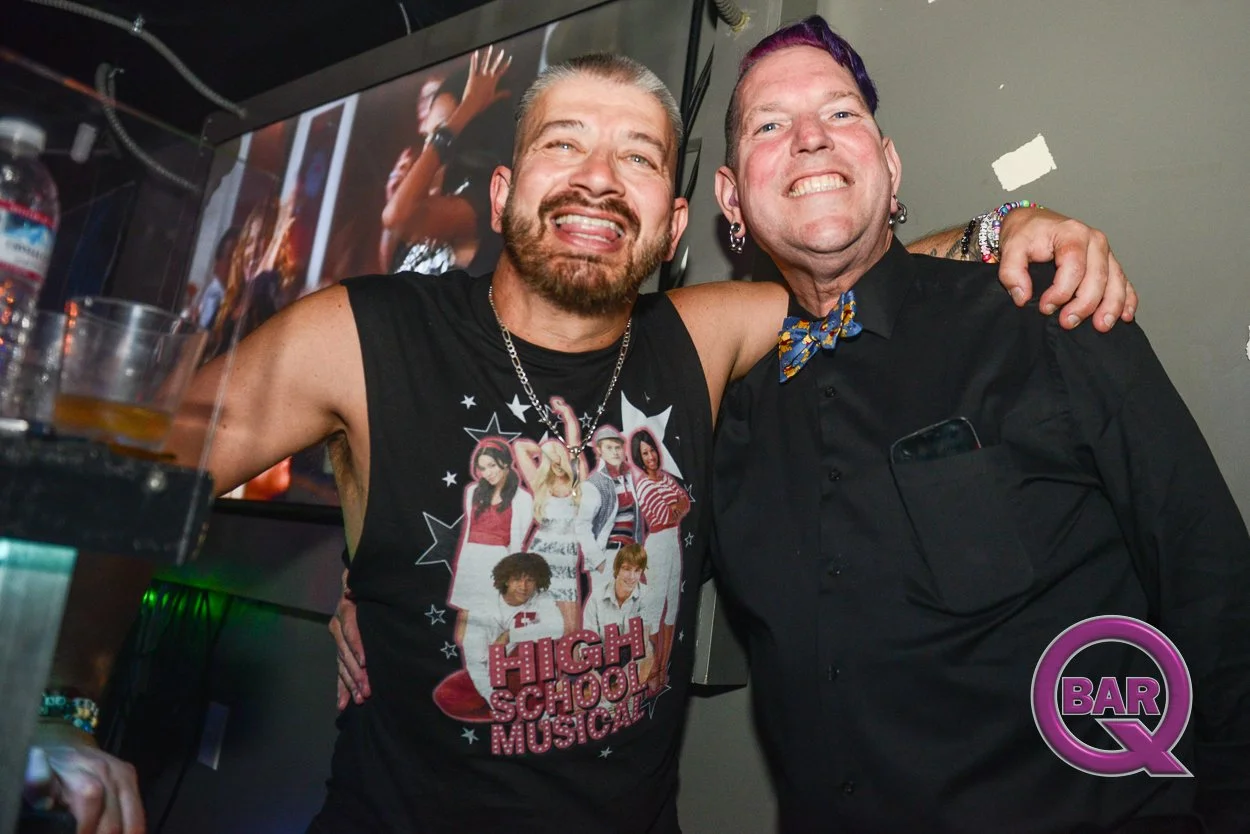 Two smiling men with arms around each other at a bar or club, one wearing a sleeveless 'High School Musical' shirt and the other in a black shirt with a colorful bow tie, in front of a TV screen.
