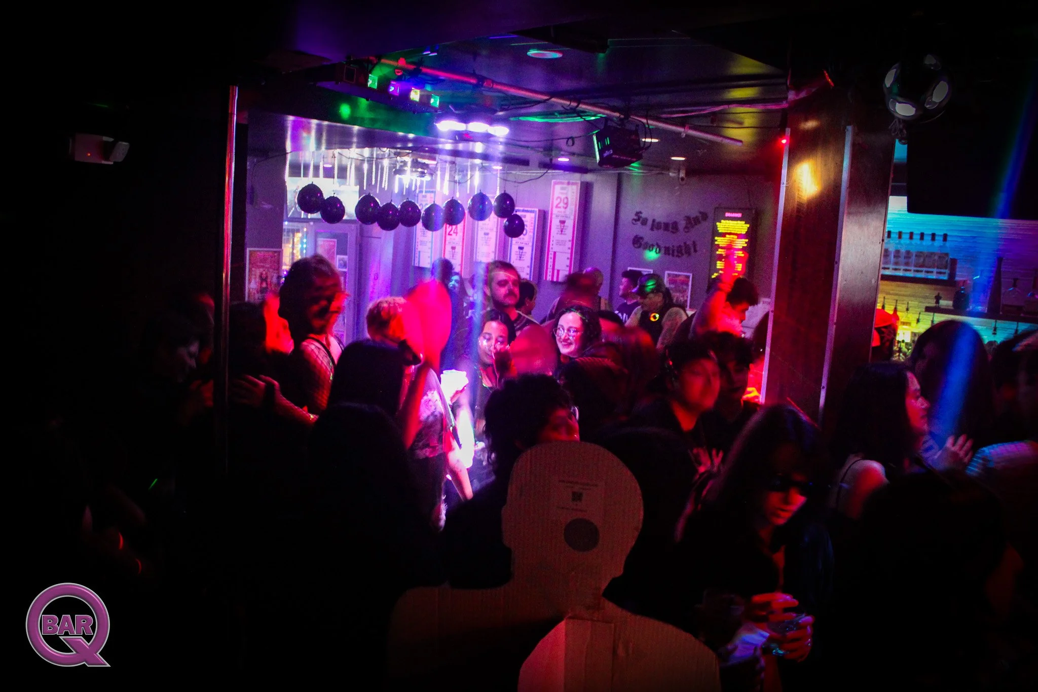 People dancing and socializing inside a nightclub with colorful lighting, balloons, and a bar area.