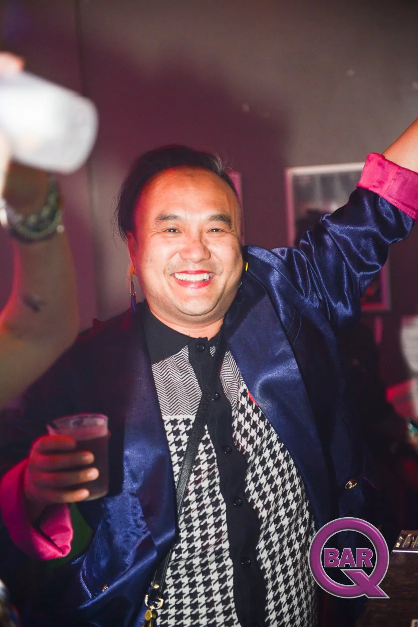 A smiling man with dark hair and earrings raising a drink in a bar environment, holding a cup with a dark beverage, with a purple 'BAR Q' logo in the bottom right corner.