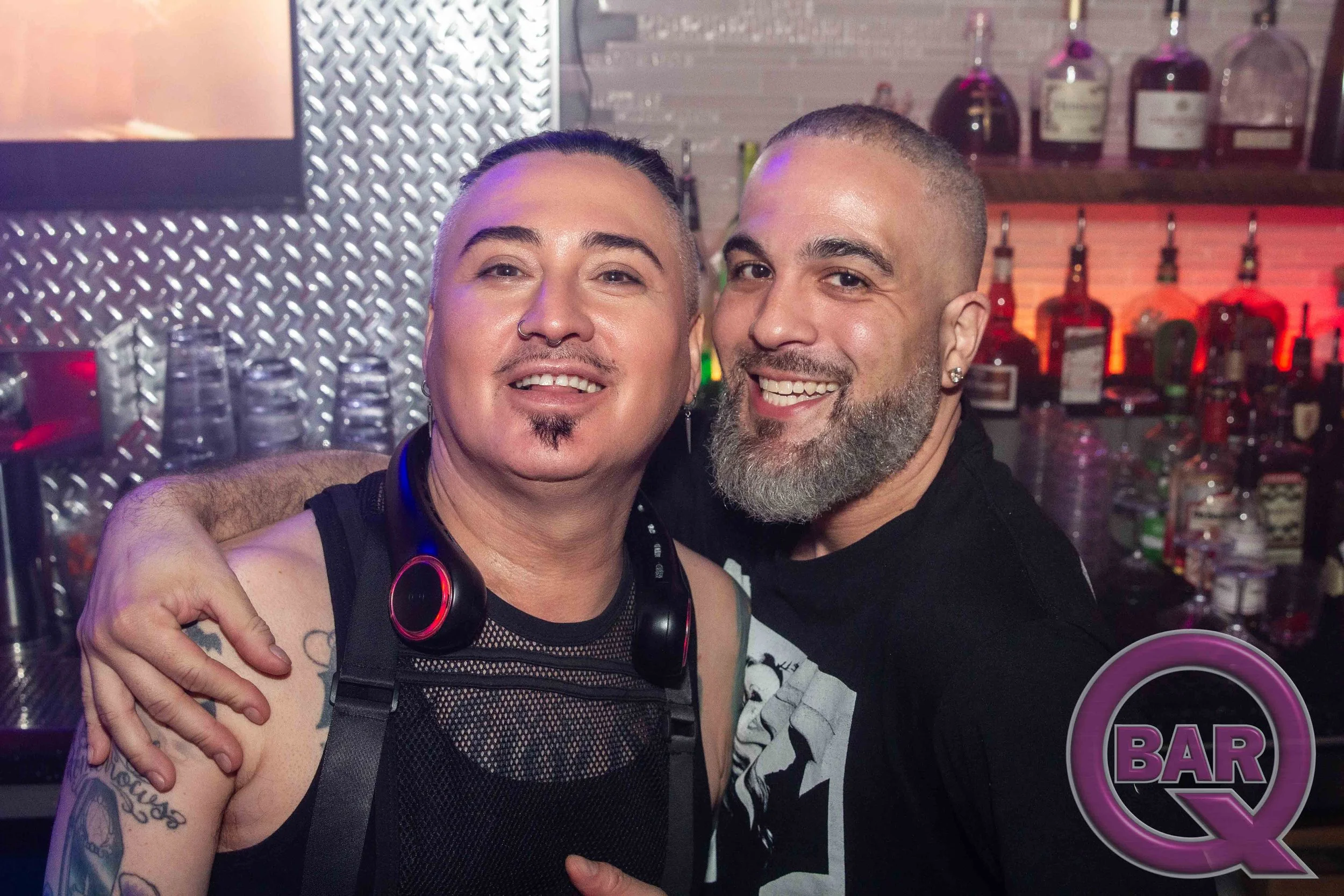 Two smiling men hugging in a bar with liquor bottles in the background.