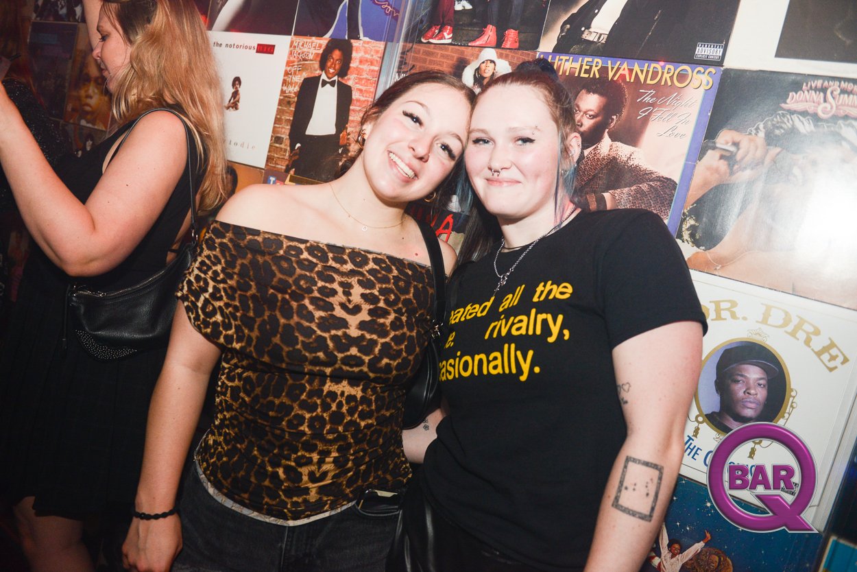 Two young women smiling at a bar, one wearing a leopard print top and the other wearing a black T-shirt with yellow text. Behind them are posters of musicians and a woman with blonde hair.