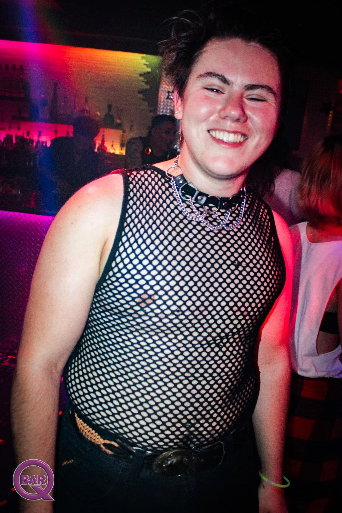 A person with short dark hair smiling in a night club or bar, wearing a fishnet top, a choker necklace, and jeans with a belt. The background has colorful lights and silhouettes of other people.
