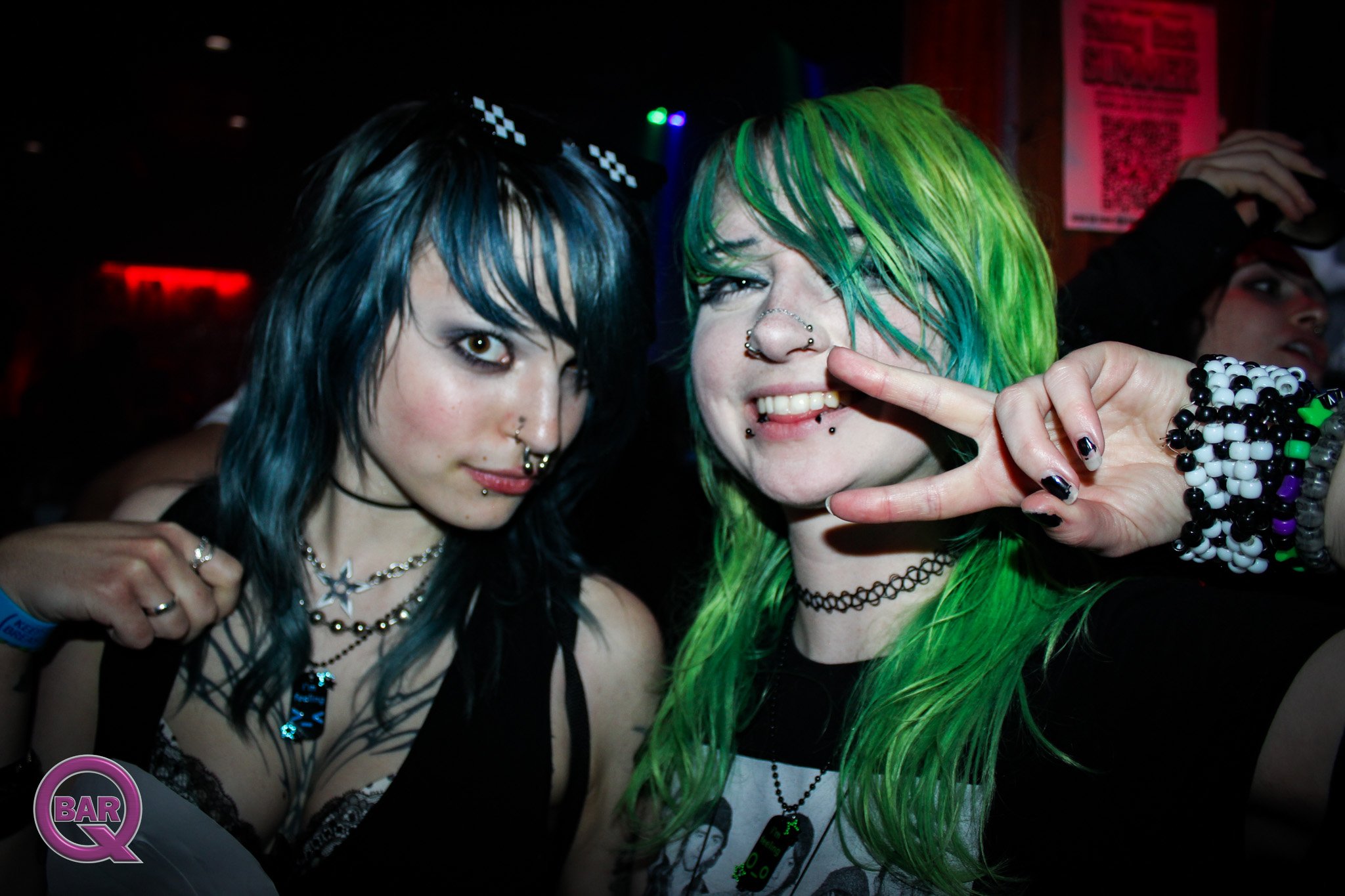 Two young women with colorful hair and piercings posing together at a nightclub. One woman has blue-black hair and the other has neon green hair. They are smiling and making playful gestures.