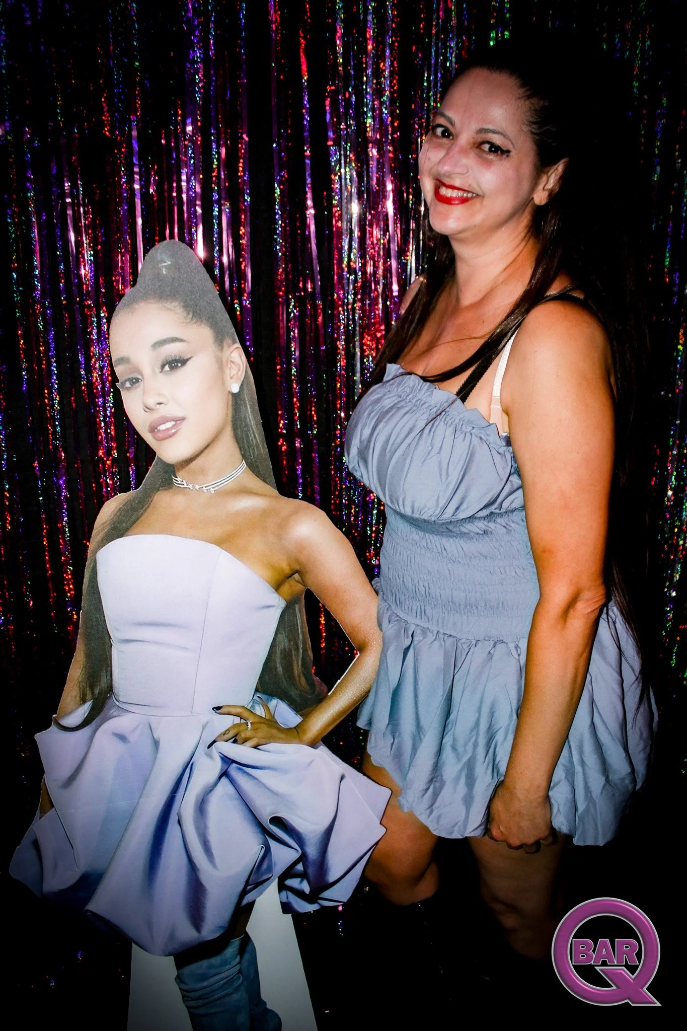 A woman in a light purple dress with black hair, standing next to a cardboard cutout of a woman in an elegant white gown with long dark hair, in front of a colorful, sparkly curtain with a purple and black 'Bar Q' logo in the bottom right corner.