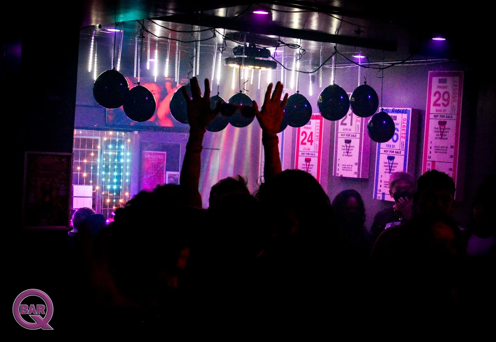 Dark nightclub scene with purple and white neon lights, hanging black balloons, hands making a peace sign, and a pink poster on the wall, with the word 'BAR' visible in the foreground.