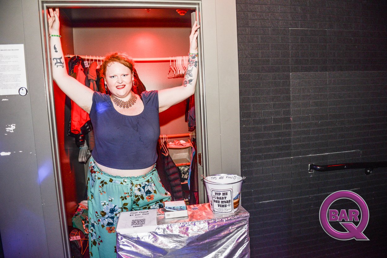 A person with red hair and tattoos inside a small elevator dressed in a navy blue crop top and floral pants, standing with arms raised, smiling at the camera, with a table and sign in front, and dark brick wall to the side. 