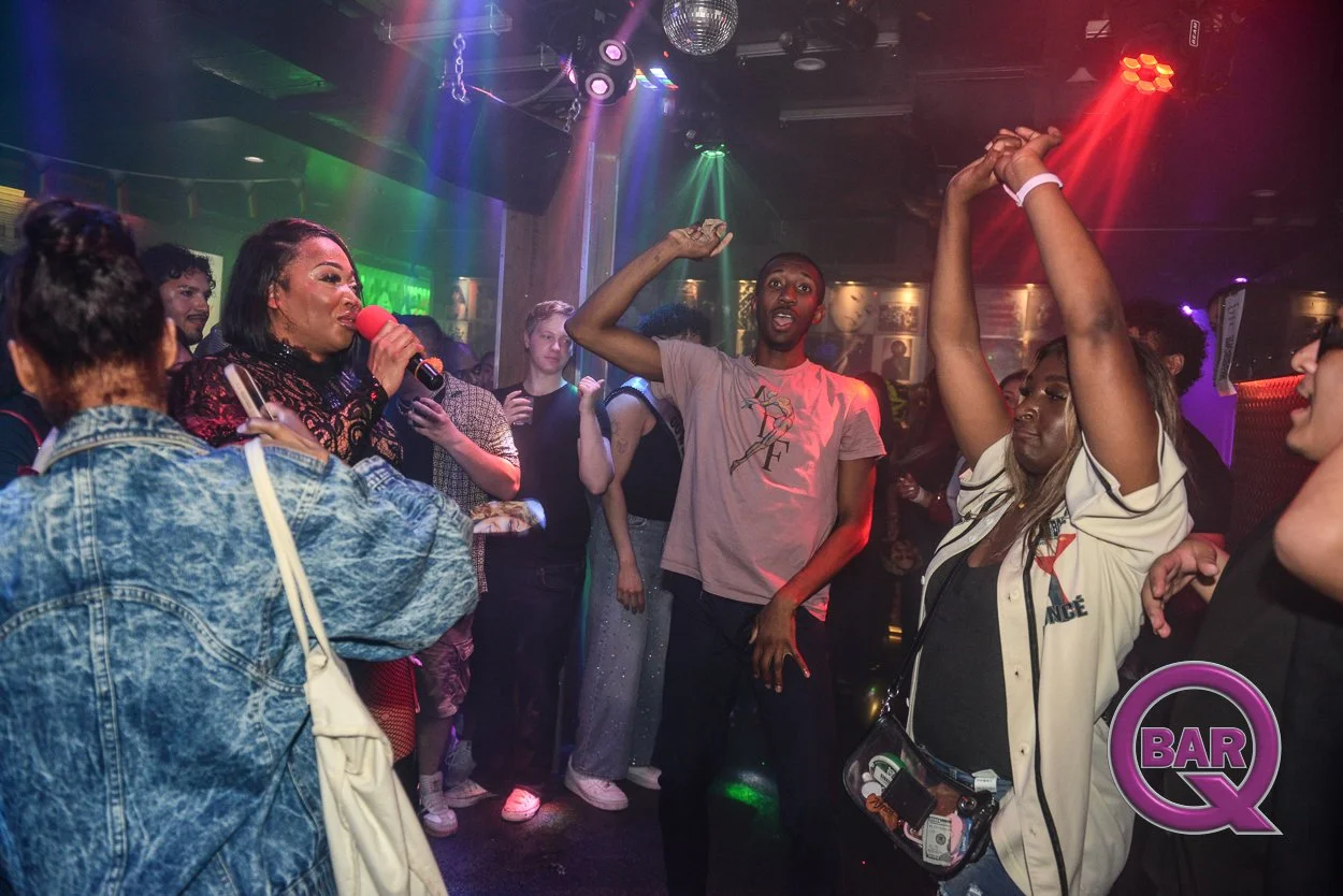 People dancing and singing in a nightclub with colorful lighting, a woman holding a microphone, and a logo that says 'Bar Q' in the corner.