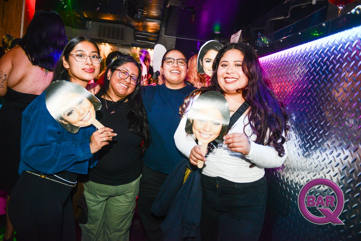 Four women at a bar holding cutout masks of a celebrity's face and smiling. Colorful lights and a metallic wall are visible in the background.