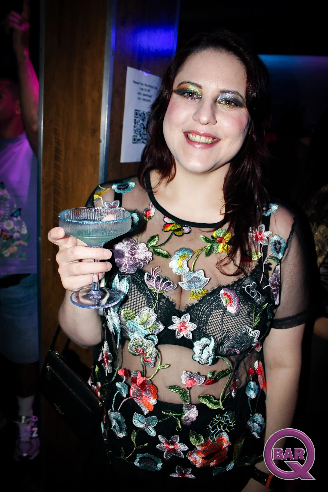 A woman with dark hair and colorful eye makeup smiling and holding a cocktail glass at a bar or club with neon lighting.
