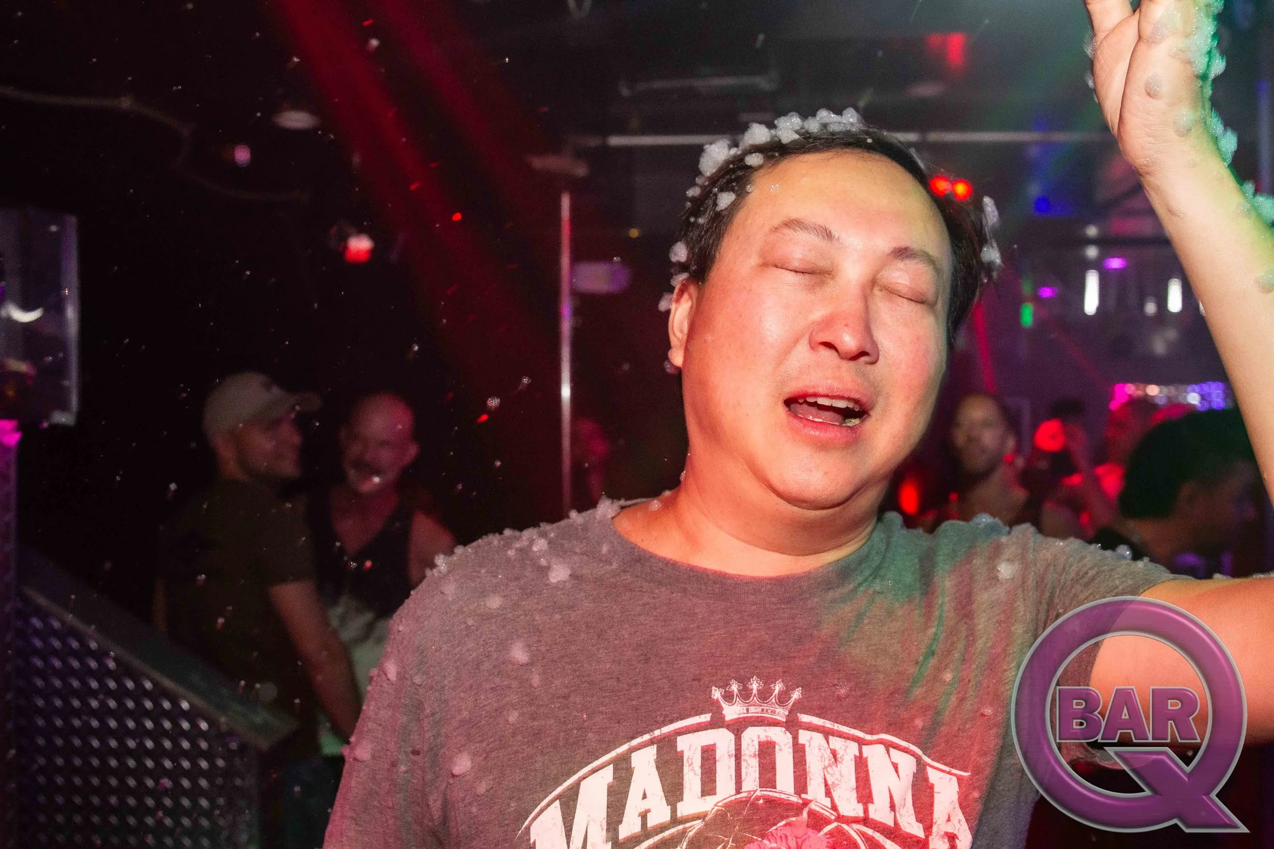 A person with short dark hair and closed eyes is enjoying foam thrown on their face at a nightclub or party. They are wearing a gray T-shirt with a graphic design. The background shows others in a dimly lit, colorful environment with bright lights an