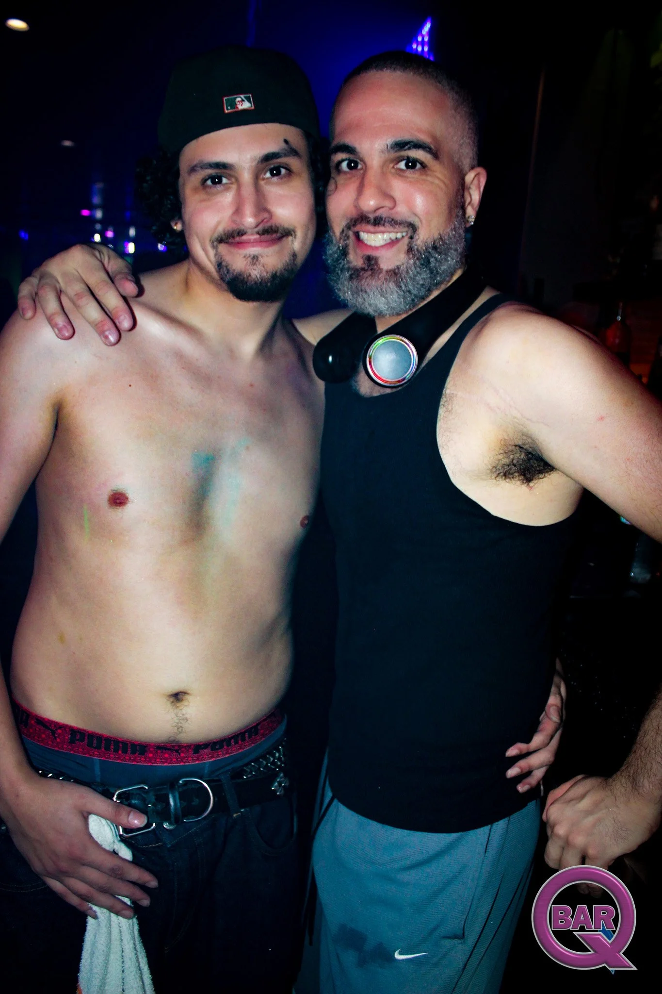 Two men smiling at a bar or nightclub with dark lighting and purple lights in the background. One man is shirtless and wearing dark jeans and a belt, the other man is wearing a black tank top with a headphone around his neck. Both men have facial hai