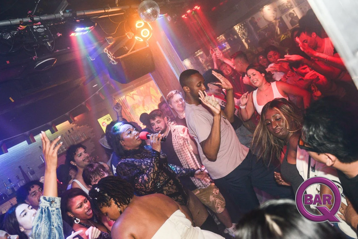 Crowd of diverse people dancing and enjoying music at a nightclub or bar, with colorful lights and a DJ booth in the background.