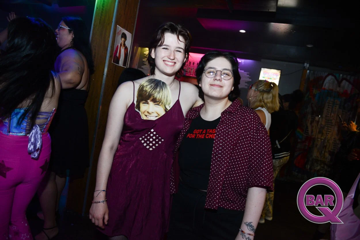 Two young women smiling at a party, with other people in the background, in a bar or club setting.