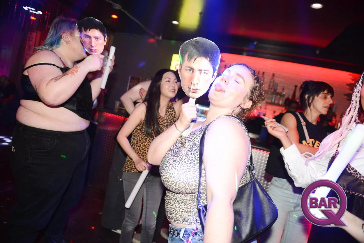 Group of people partying in a nightclub, some holding oversized posters of a man's face, with colorful lighting and a bar in the background.