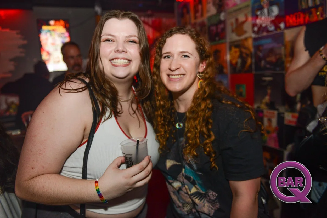 Two women smiling and posing together at a bar, one holding a drink, in a lively, colorful setting with posters on the wall.