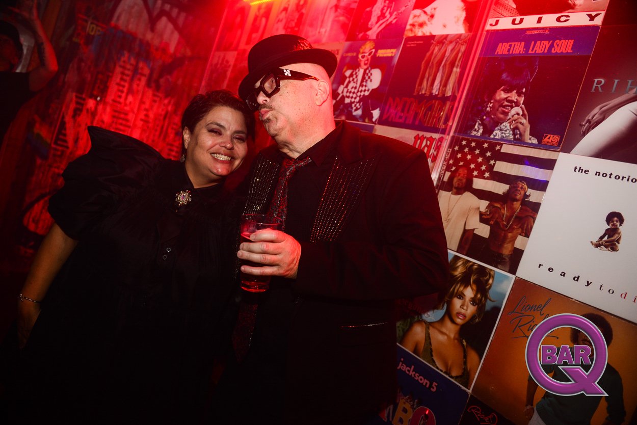 A man wearing glasses, a bowler hat, and a black suit with red accents is holding a drink and leaning toward a woman in a black dress with puffed sleeves, both smiling at a party or club. The background is decorated with a collage of album covers and